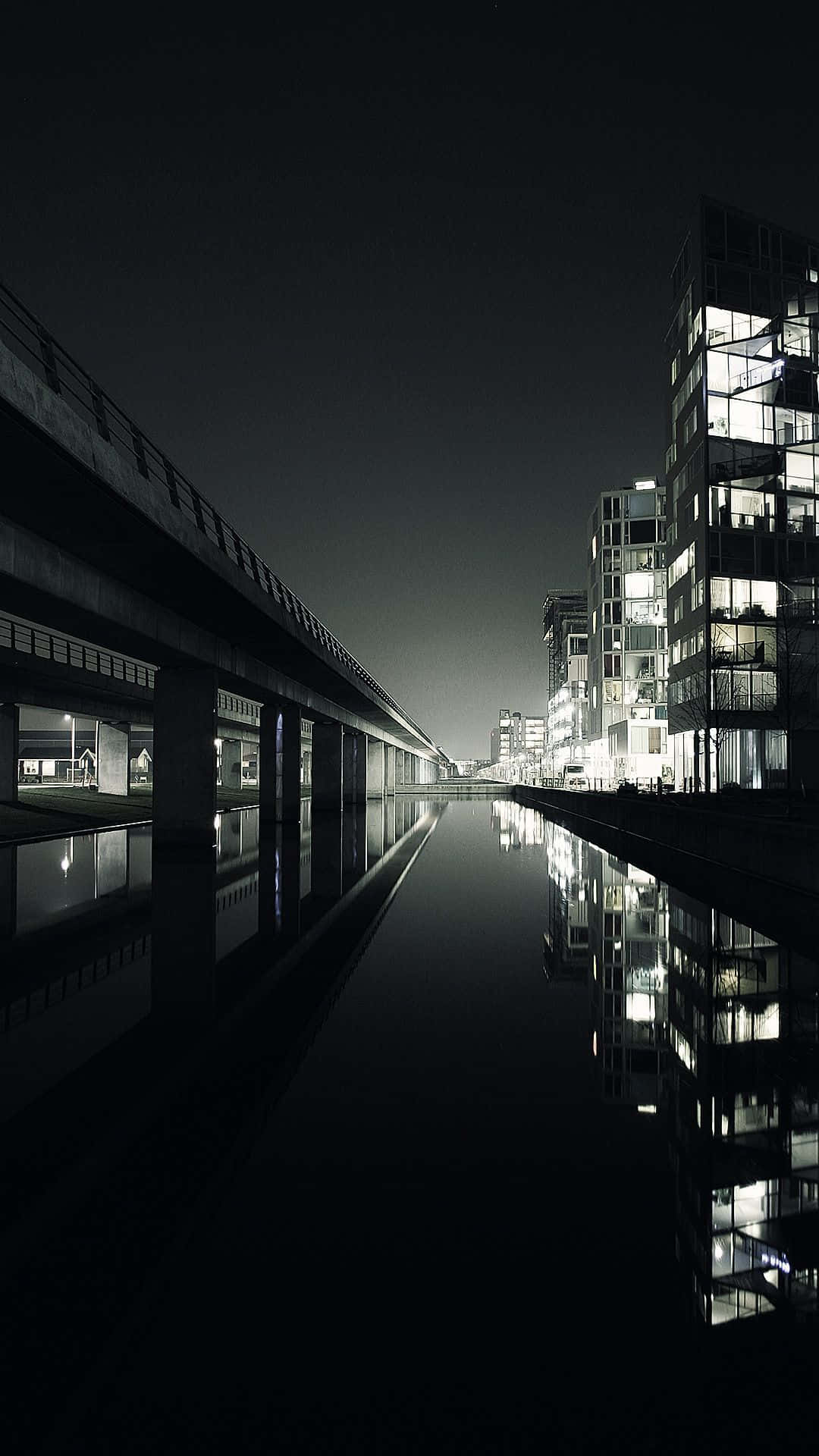 黑暗城市 桌布 壁紙 — 一個嚴峻的夜景城市景觀，發光的窗戶倒映在混凝土天橋下的平靜運河中。