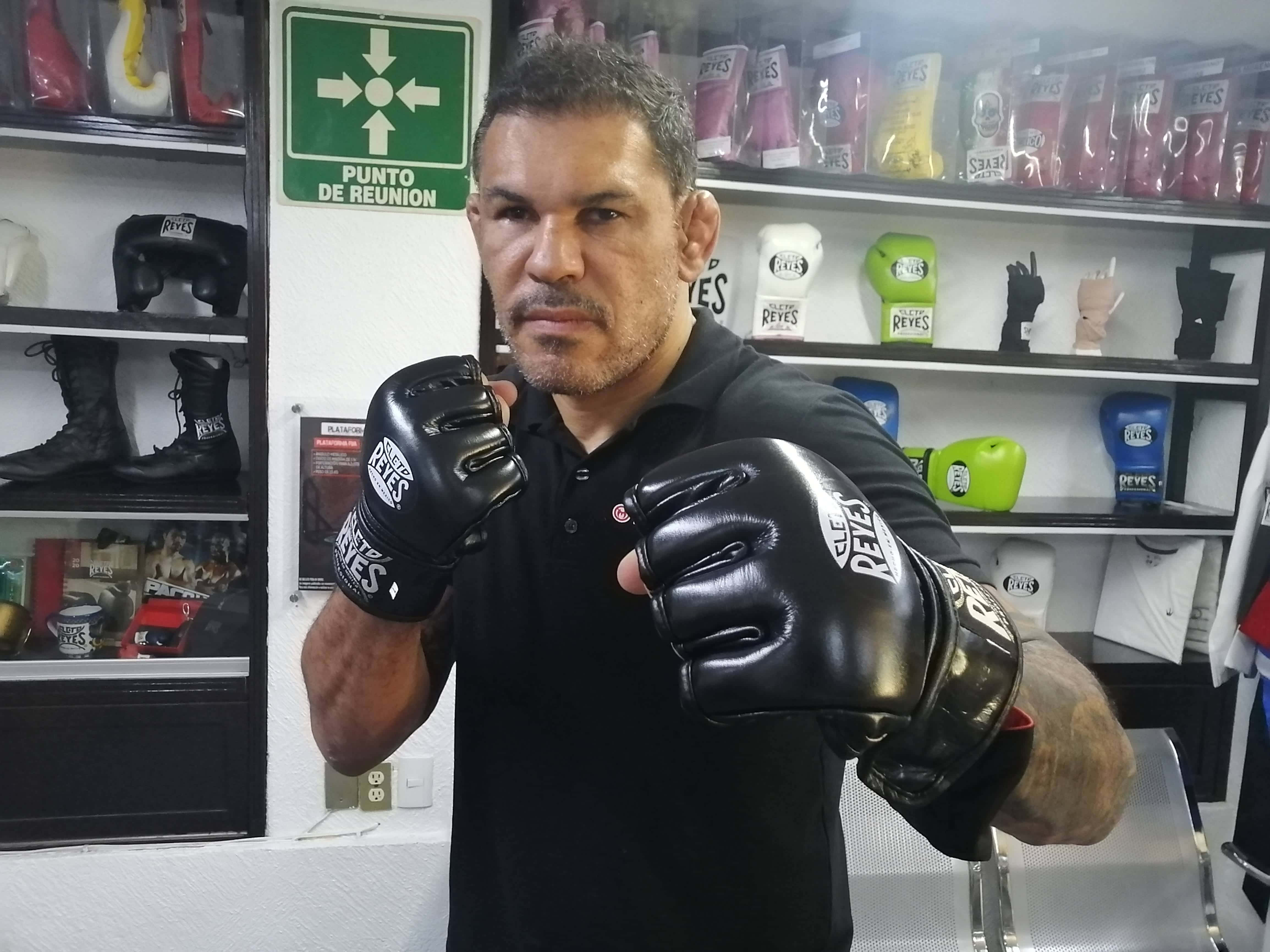 MMA 桌布 壁紙 — 一個嚴肅的拳擊手戴著黑色手套，站在被拳擊設備和品牌裝備包圍的健身房中。