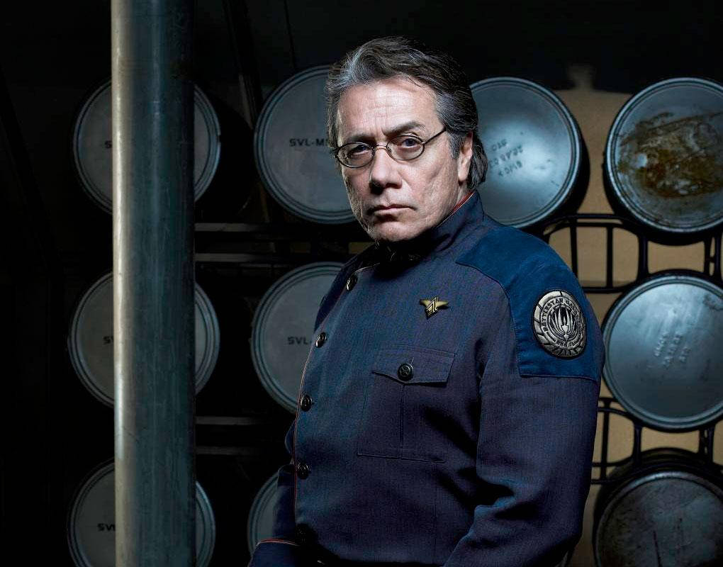 actor 桌布 壁紙 — 一個嚴肅的男人，身穿帶有徽章的深藍色制服，站在一個光線昏暗的工業空間，周圍是金屬桶。