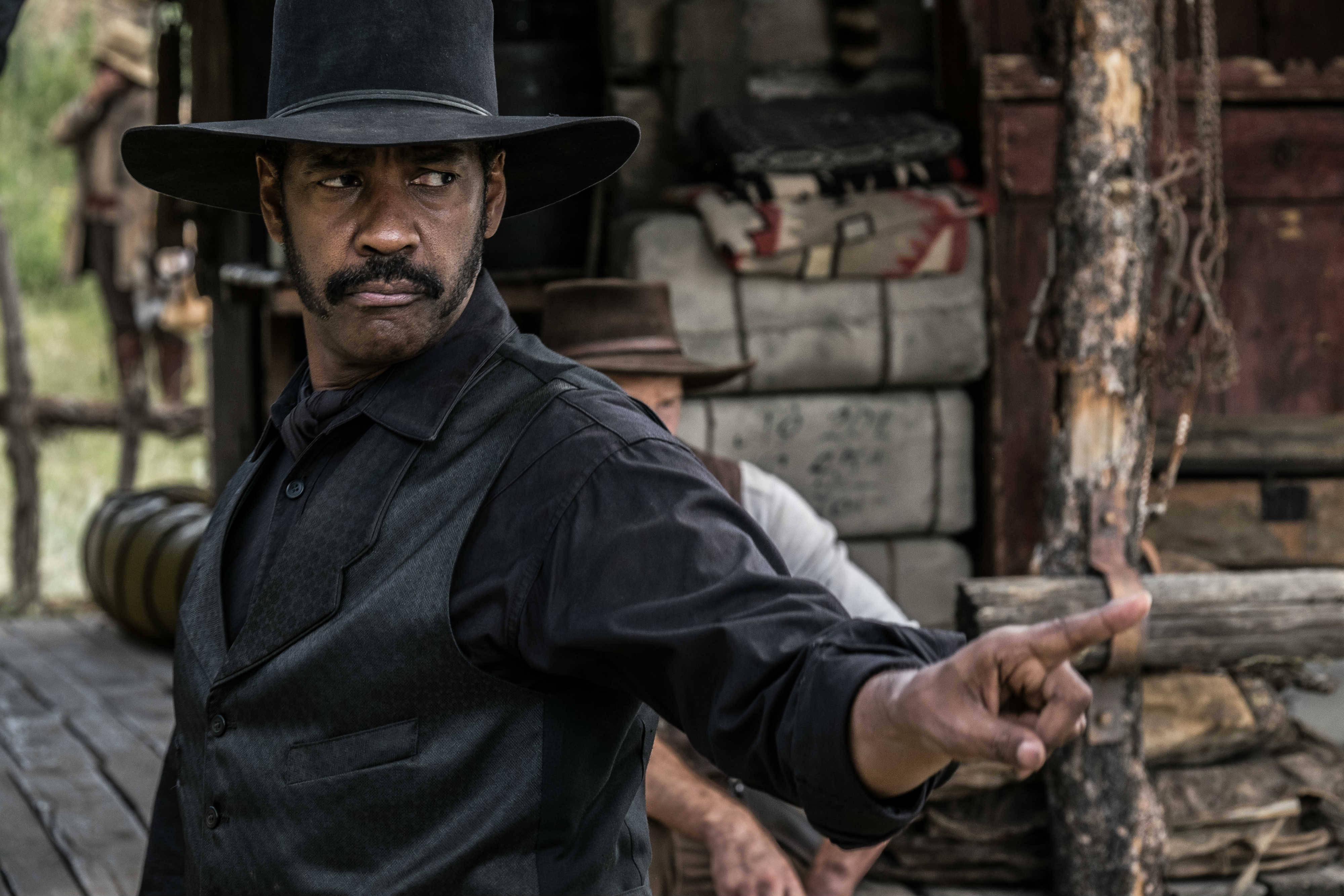 Denzel Washington 桌布 壁紙 — 一個嚴肅的男人戴著黑帽和背心，在質樸的木製邊疆場景中充滿強度地指向前方。