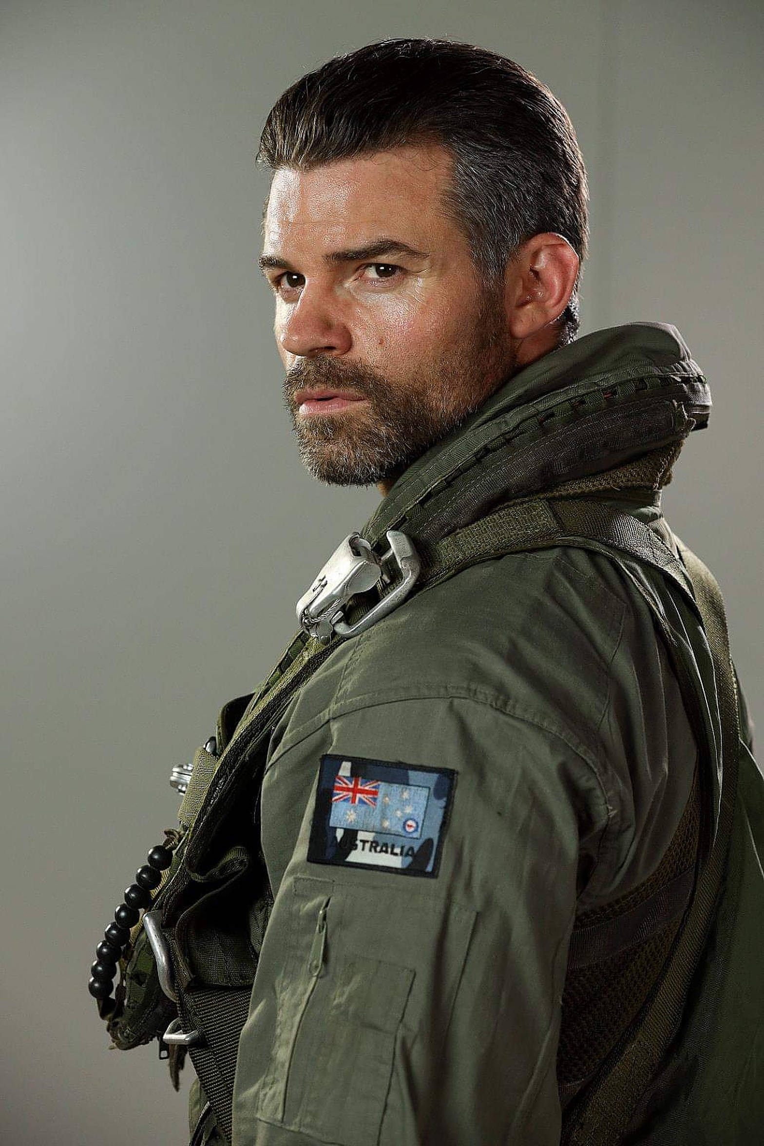 Daniel Gillies 桌布 壁紙 — 一個嚴肅的男人穿著橄欖綠色的軍用飛行夾克，帶有澳洲徽章，在純灰色背景上專注地越過肩膀凝視。