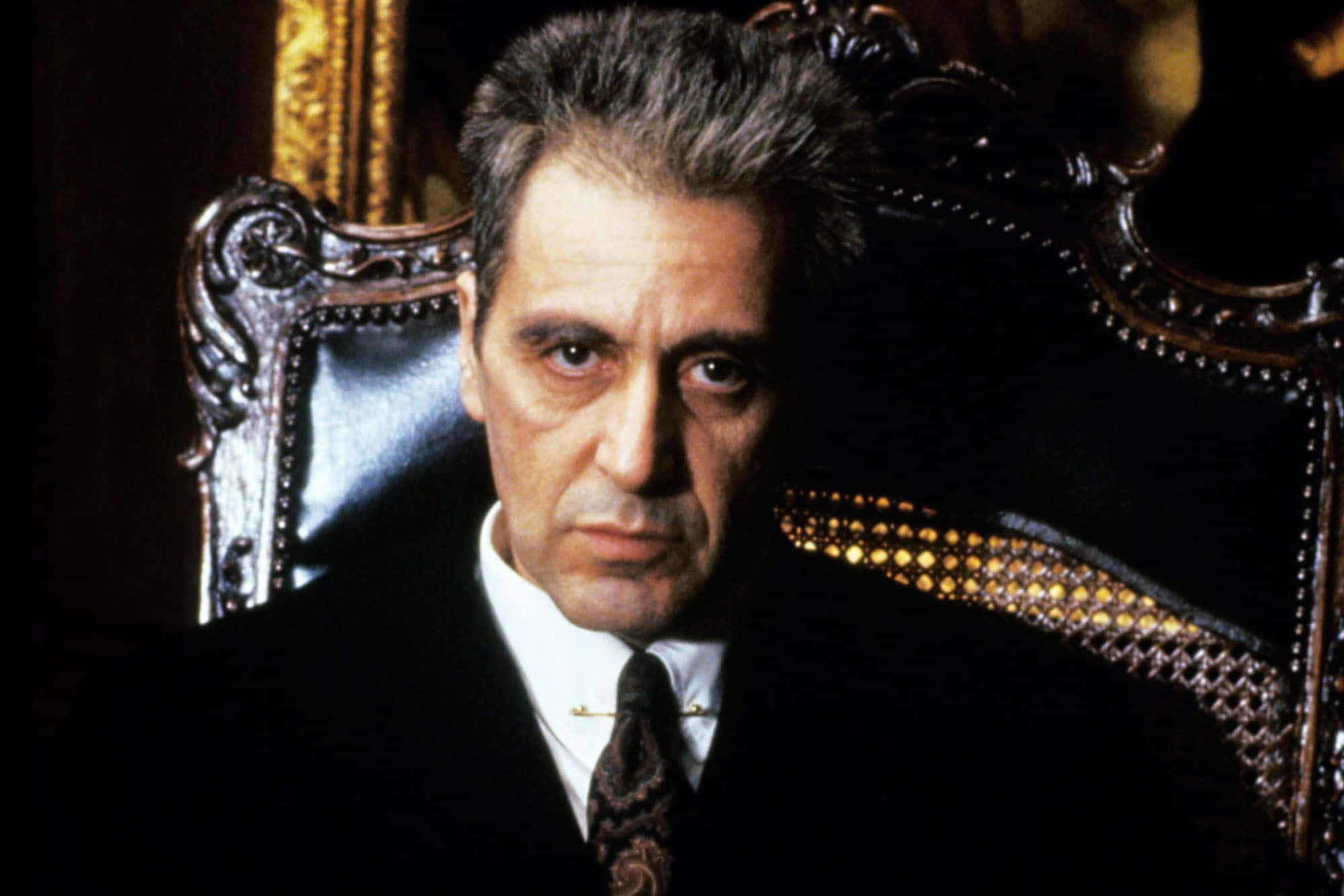 Michael Corleone 桌布 壁紙 — 一個嚴肅的男人穿著深色西裝，坐在華麗的椅子上，在戲劇性的燈光下帶著強烈的目光。