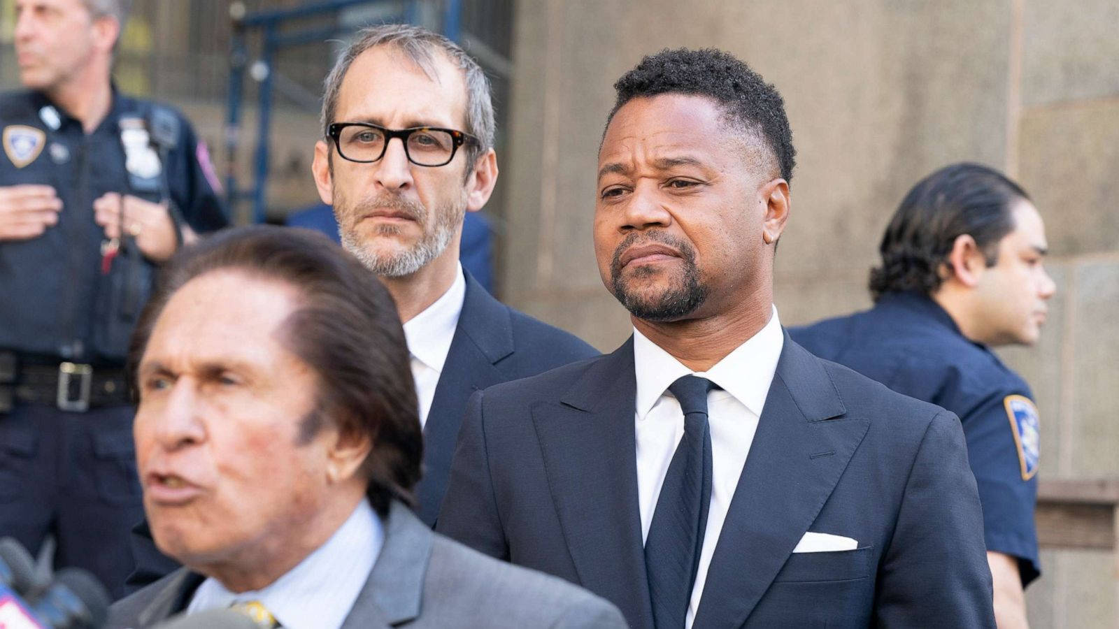 Celebrity 桌布 壁紙 — 一個嚴肅的男人穿著深色西裝站在戶外，兩旁有警察和另一個穿西裝的男人。