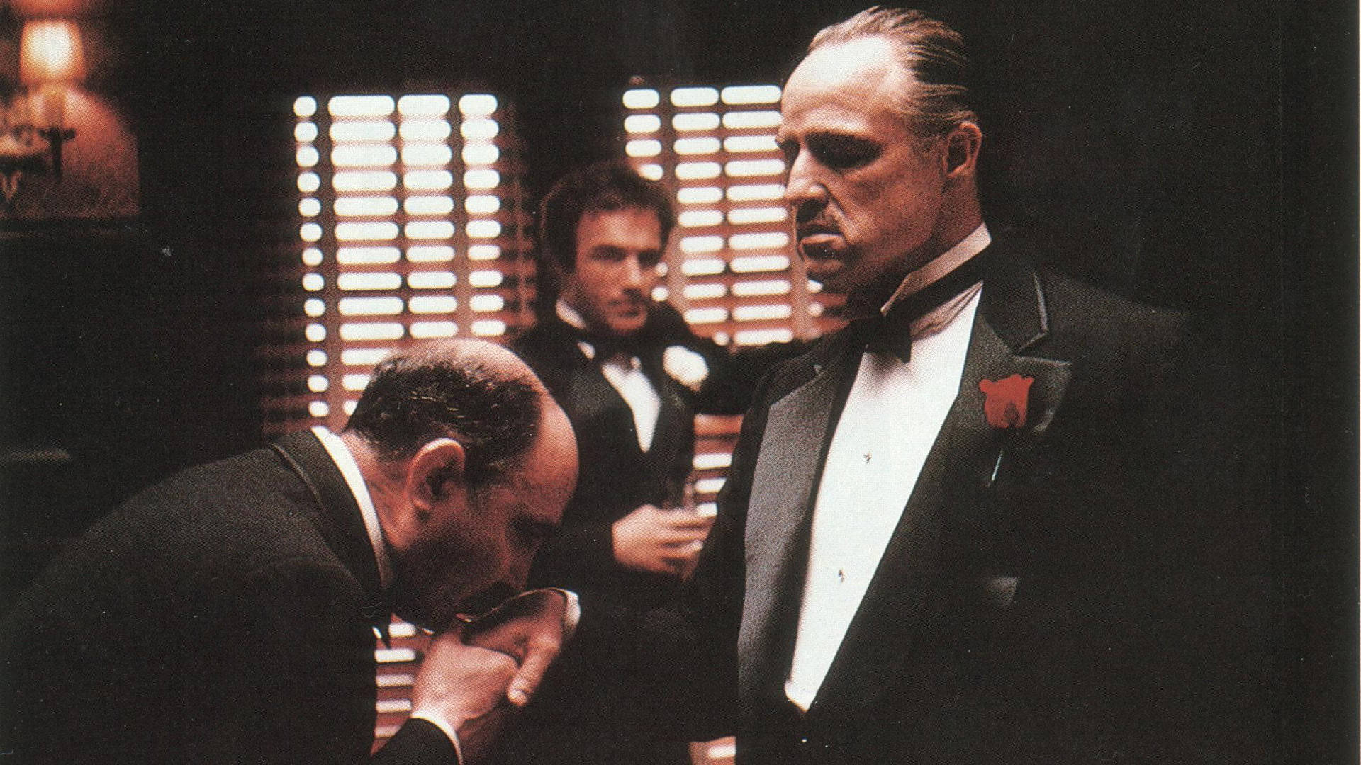 Godfather 桌布 壁紙 — 一個嚴肅的男人穿著燕尾服，別著紅玫瑰胸針，站在另一個在昏暗房間裡深深鞠躬的男人上方，房間有百葉窗。