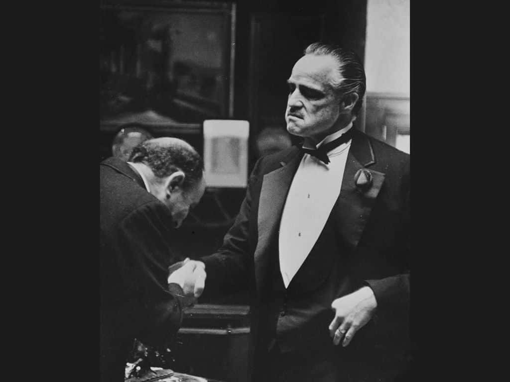 Michael Corleone 桌布 壁紙 — 一個嚴肅的男人穿著燕尾服，在昏暗的房間裡將手放在另一個男人的肩膀上。