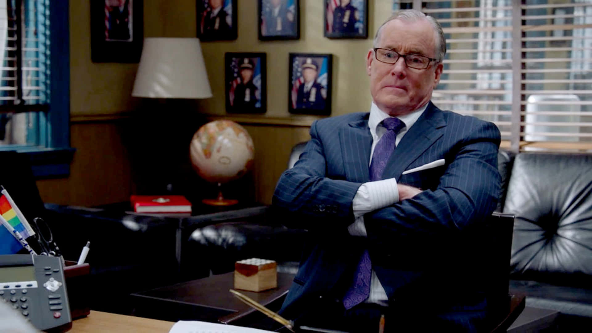 John C. Mcginley 桌布 壁紙 — 一個嚴肅的男人穿著細條紋西裝，雙臂交叉坐在皮椅上，在一個燈光昏暗的辦公室裡，裝飾著裱框照片和一個地球儀。