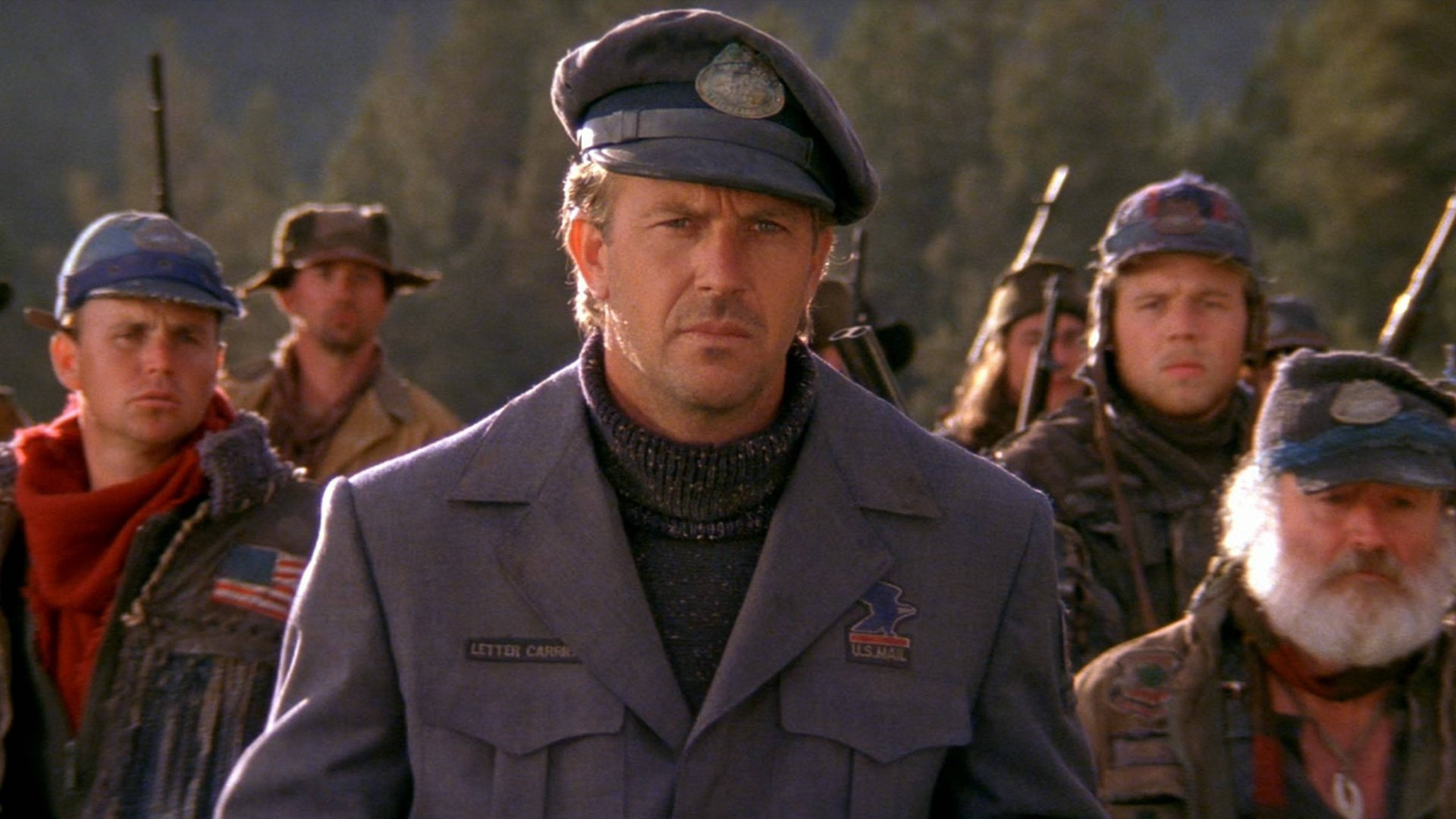 Kevin Costner 桌布 壁紙 — 一個嚴肅的男人穿著藍色制服和帽子站在畫面中央，身後有武裝男子穿著粗獷裝備，背景是森林山區。