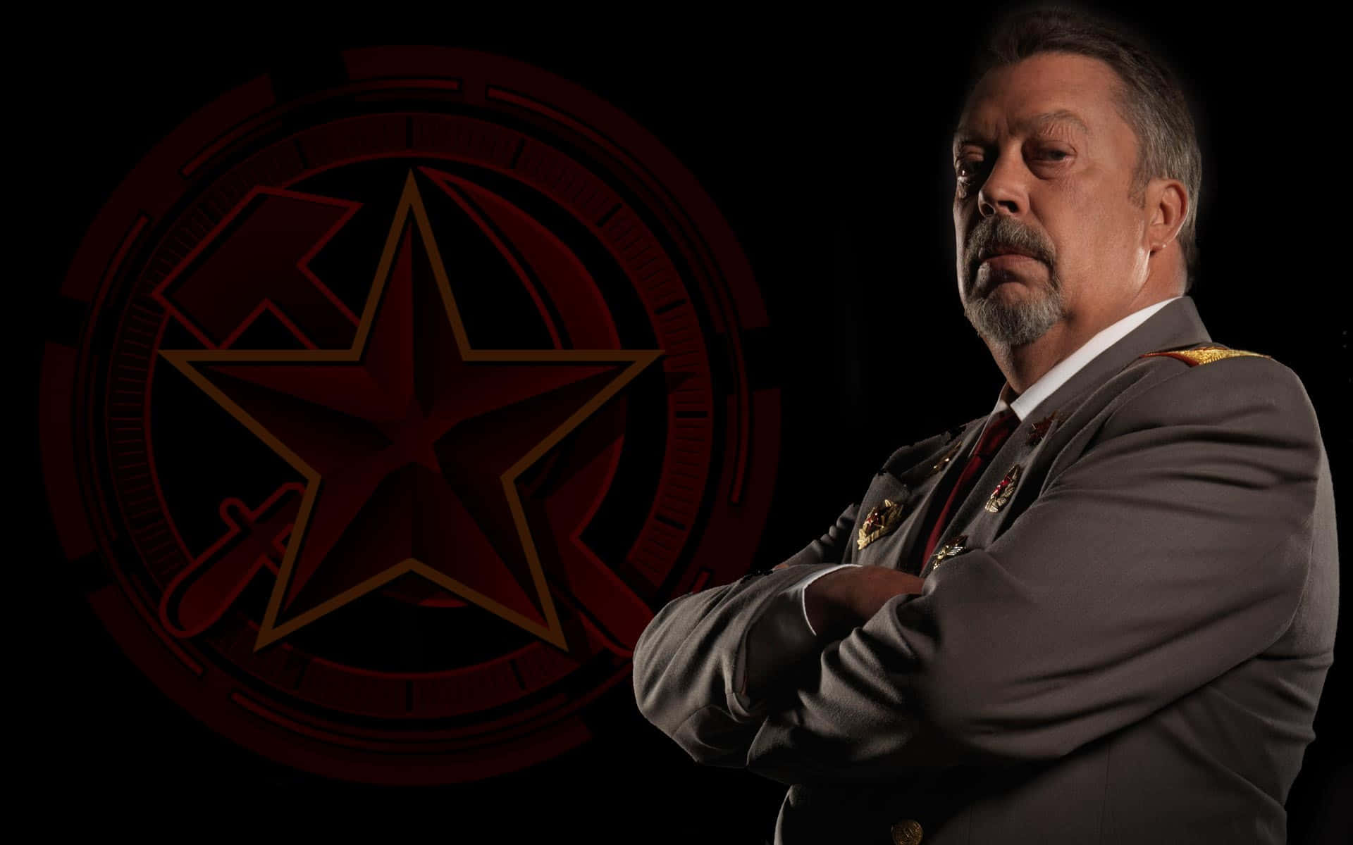 Tim Curry 桌布 壁紙 — 一個嚴肅的男人穿著軍服，雙手交叉站在一個黑暗背景前面，背景有一個發光的紅色星星標誌。