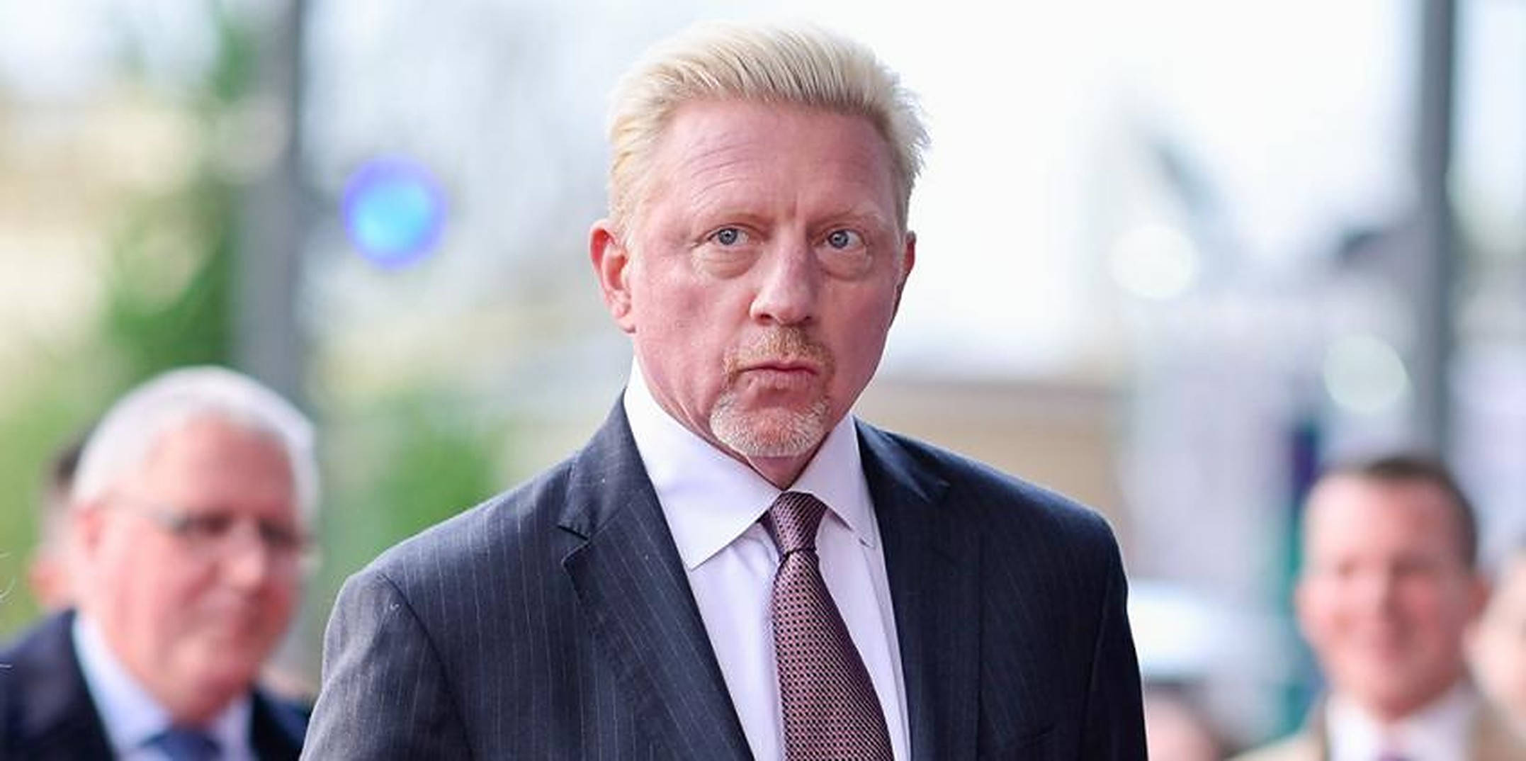 Boris Becker 桌布 壁紙 — 一個嚴肅的金髮男子穿著深色細條紋西裝和圖案領帶，站在戶外，身後有模糊的人群。