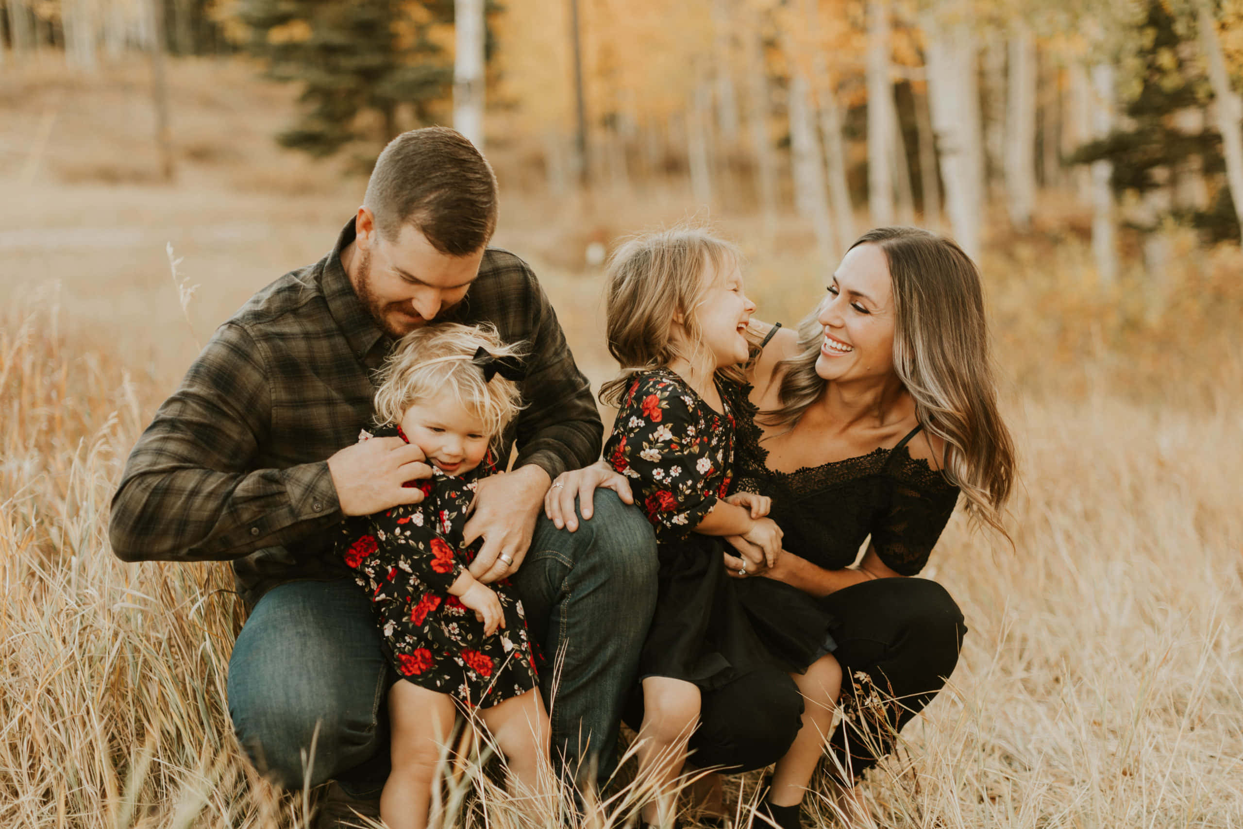 Fall Family 桌布 壁紙 — 一個四口之家微笑著蹲在秋天樹下的金色高草中，分享歡樂的笑聲。
