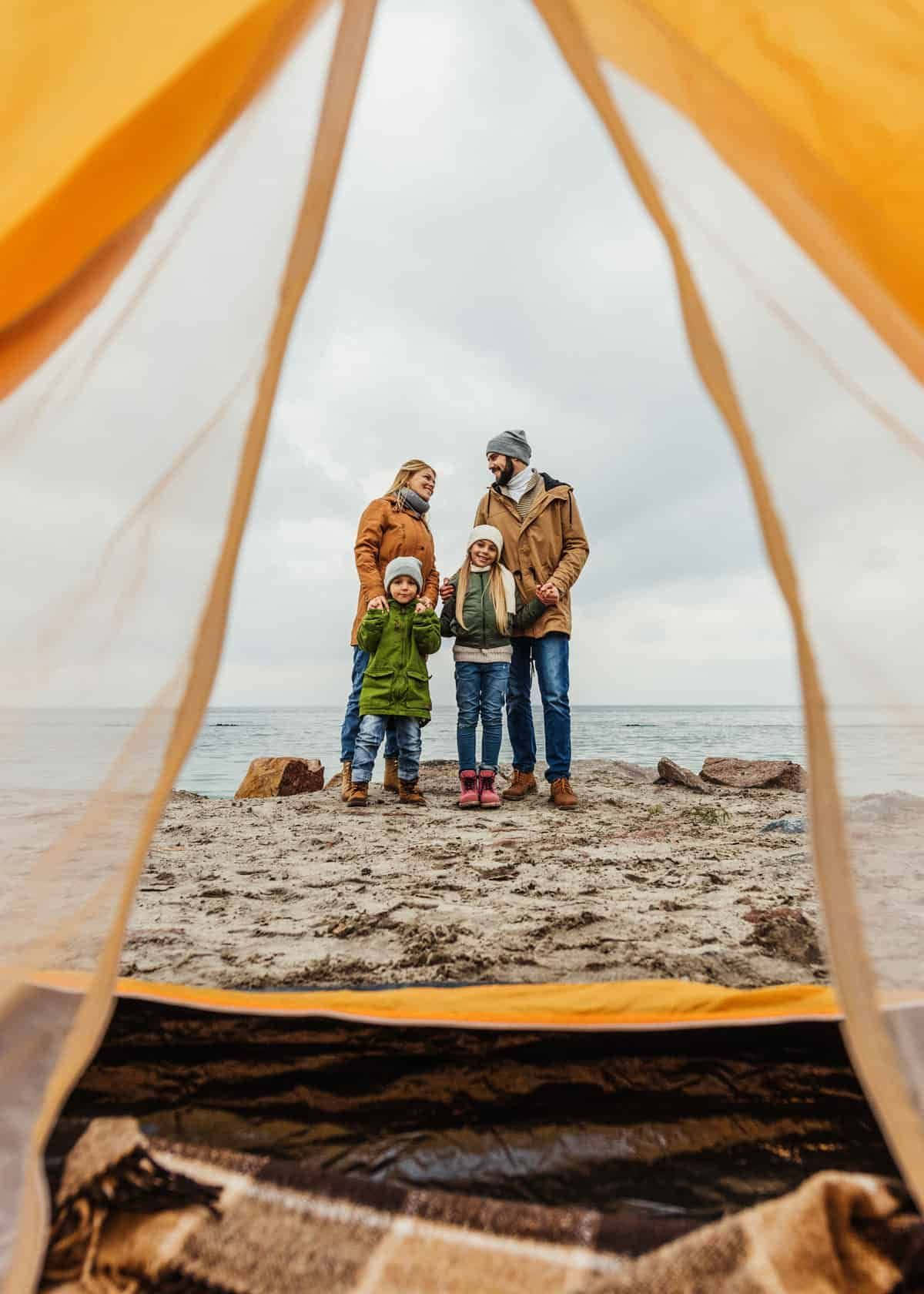 Minimalist Camping 桌布 壁紙 — 一個四口之家站在岩石海灘上，透過帳篷的橙色布料看到。