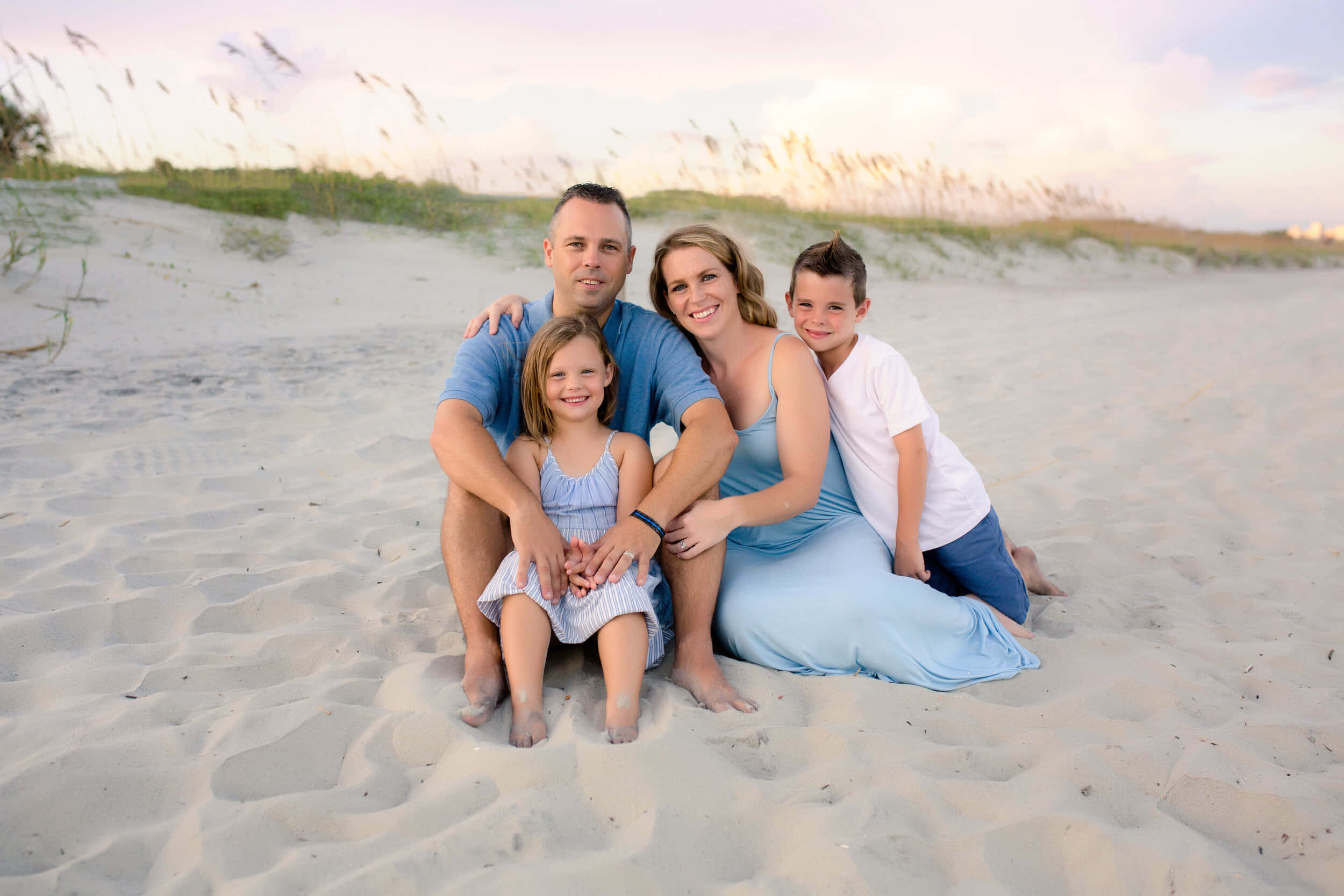 Beach Family 桌布 壁紙 — 一個四口之家面帶微笑，在金色日落時分的沙灘上合照，背後有沙丘和草叢。