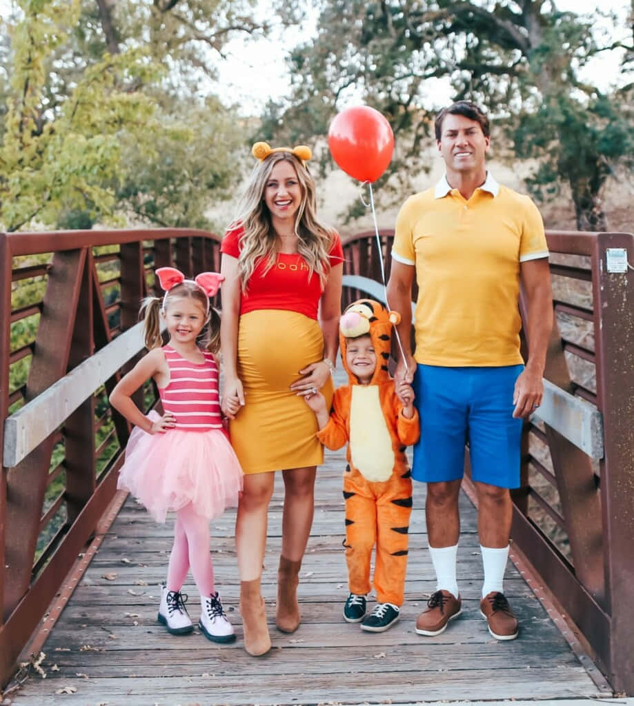 Family 桌布 壁紙 — 一個四口之家面帶微笑，穿著小熊維尼主題服裝，在樹木環繞的木橋上擺姿勢。
