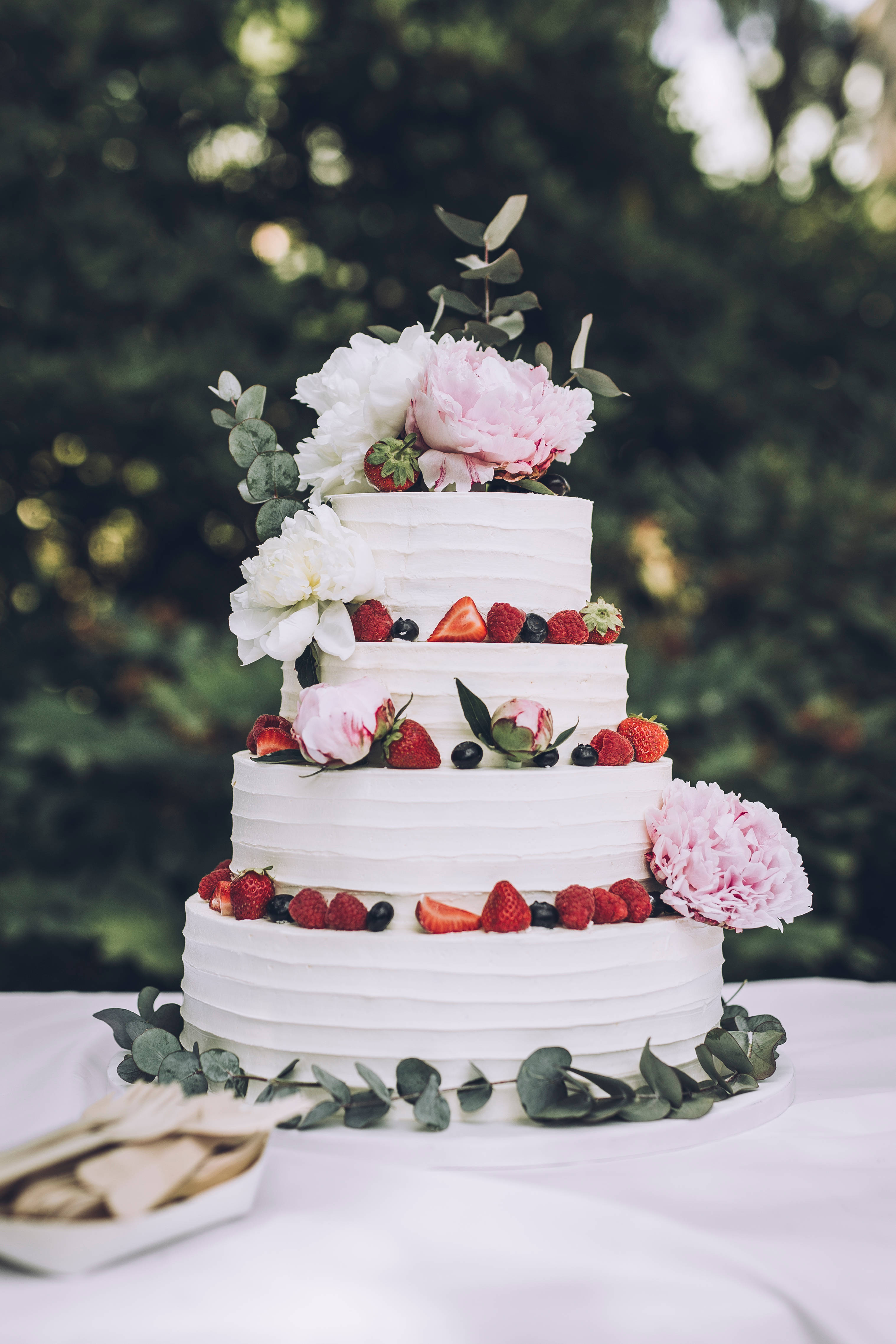 Wedding 桌布 壁紙 — 一個四層白色婚禮蛋糕，飾有粉紅色牡丹、草莓和藍莓，優雅地放在戶外的白色桌布上。
