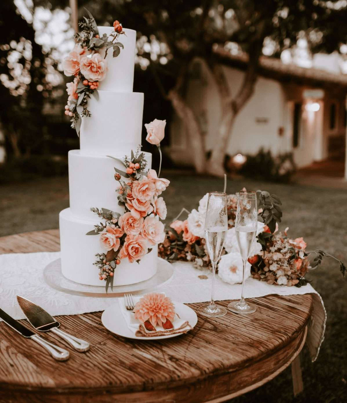 Wedding 桌布 壁紙 — 一個四層白色婚禮蛋糕，飾有蜜桃玫瑰和綠葉，優雅地放置在戶外的鄉村風格木桌上。