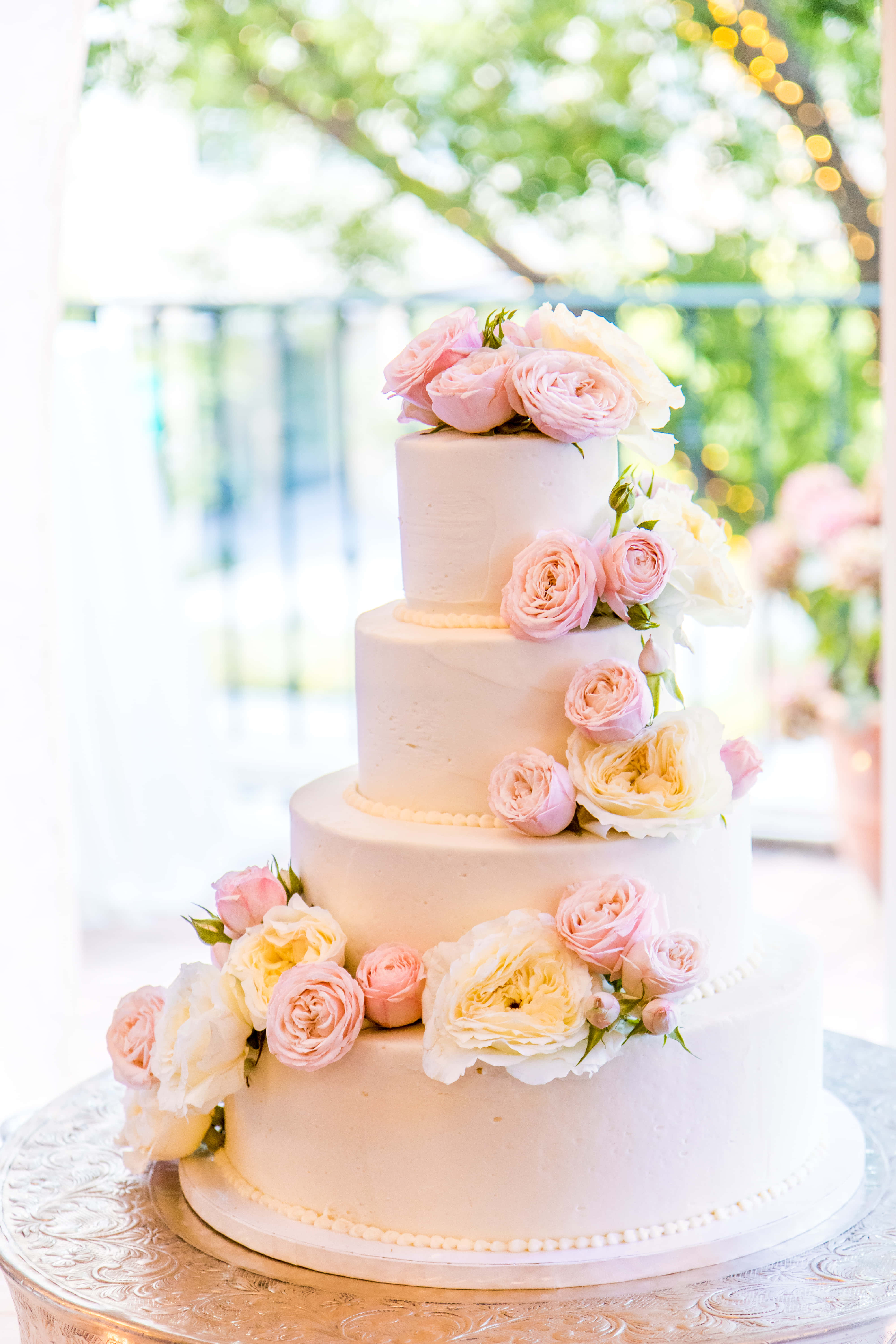 Cake Fondant 桌布 壁紙 — 一個四層白色結婚蛋糕，飾以柔和粉紅和奶油色玫瑰，優雅地放在戶外的銀色架上。