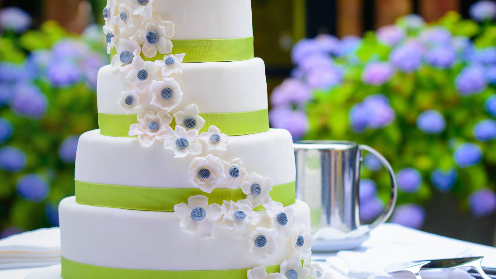Wedding 桌布 壁紙 — 一個四層白色結婚蛋糕，飾以萊姆綠色絲帶和層疊的白色糖花，放在戶外一個銀色馬克杯旁邊。