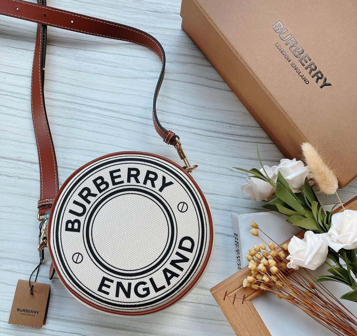 Burberry 桌布 壁紙 — 一個圓形 Burberry England 斜背包，配有棕色皮帶，放在白色木質表面上，旁邊是其盒子和白色花卉佈置。