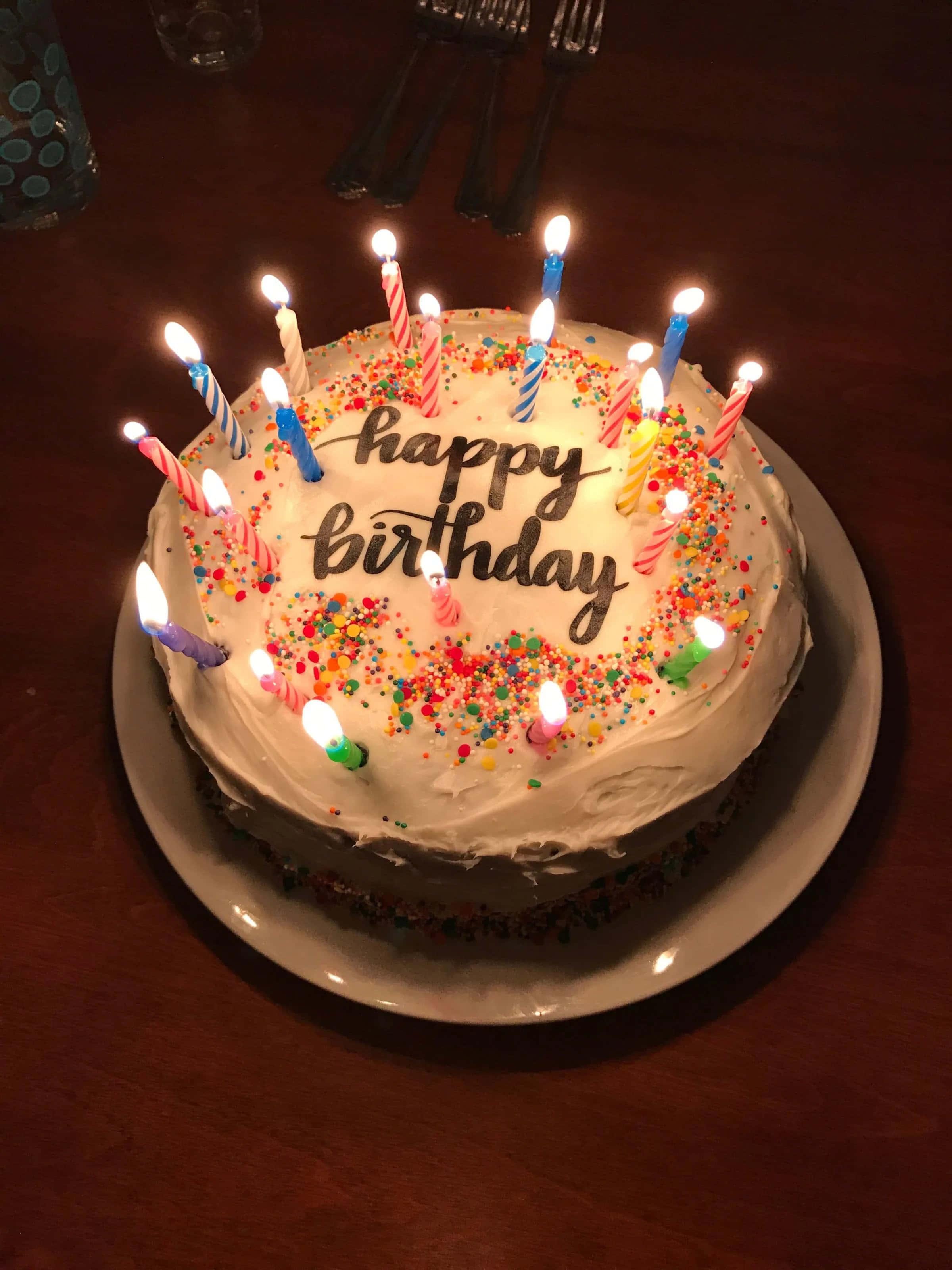 Happy Birthday Cake 桌布 壁紙 — 一個圓形生日蛋糕，上面有燃燒的蠟燭和彩色糖粒，放在木桌上，在溫暖的室內燈光下。