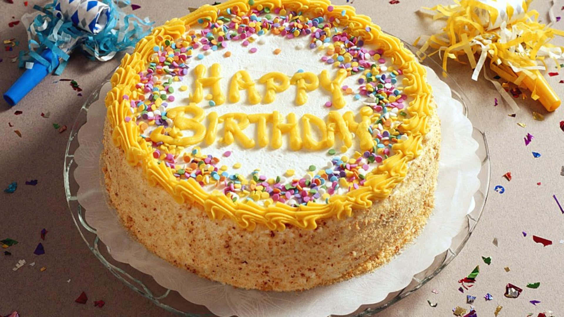 蛋糕 桌布 壁紙 — 一個圓形生日蛋糕，上面有黃色糖霜和彩色糖粒，放在玻璃盤上，周圍散落著五彩紙屑和派對喇叭。