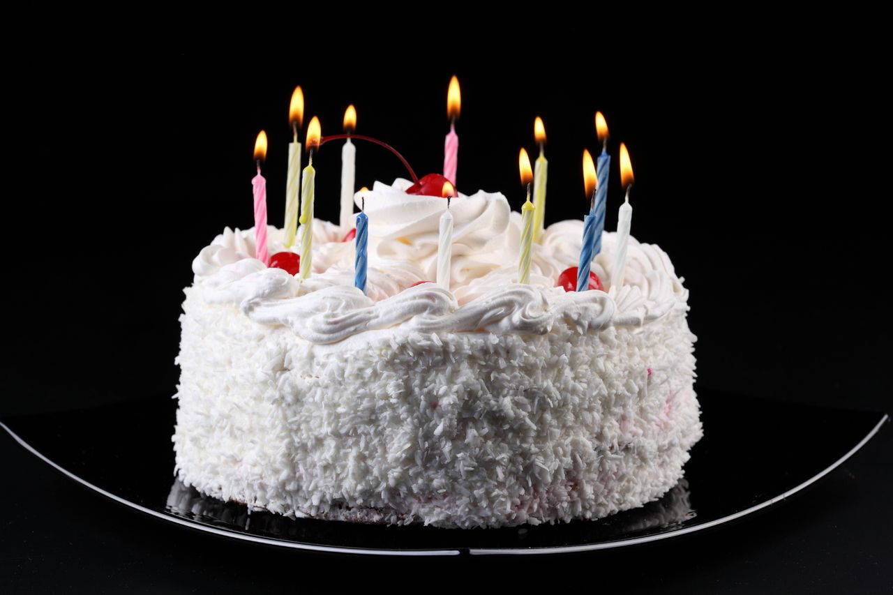Birthday Cake 桌布 壁紙 — 一個圓形白色椰子蛋糕，上面有燃燒的彩色蠟燭和櫻桃，放在黑色盤子上，背景深色。