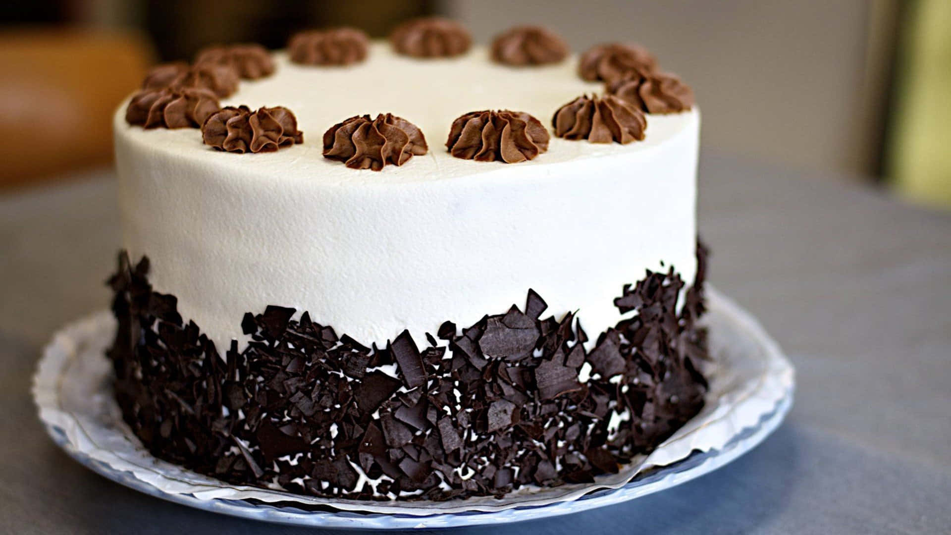 Black Forest Cake 桌布 壁紙 — 一個圓形白色糖霜蛋糕，頂部飾有巧克力漩渦，底部周圍有巧克力碎屑，優雅地放在盤子上。
