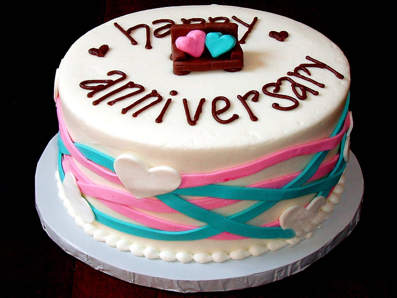 Happy Anniversary 桌布 壁紙 — 一個圓形白色週年蛋糕，裝飾有粉紅色和藍綠色絲帶漩渦、巧克力心形和「happy anniversary」字樣。