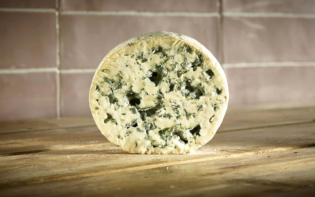 Blue Cheese 桌布 壁紙 — 一個圓形的藍紋起司輪，帶有綠色紋理，直立於質樸的木質表面上，背景是磁磚牆。