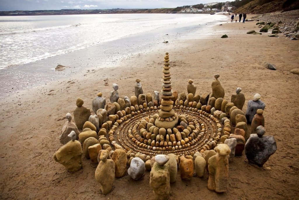 Land Art 桌布 壁紙 — 一個圓形石雕，中央有一座塔，矗立在濕漉漉的沙灘上，海浪翻滾而來，遠處的人們沿著海岸漫步。