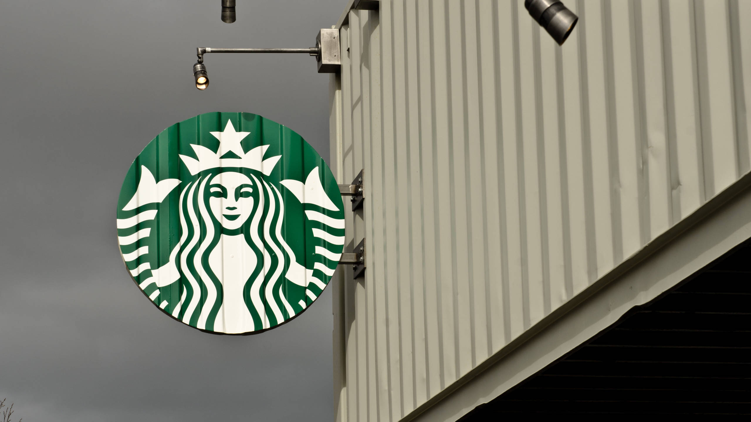 星巴克 桌布 壁紙 — 一個圓形綠色Starbucks標誌，上面有人魚和白色星星，掛在灰色天空下的波紋金屬建築上。