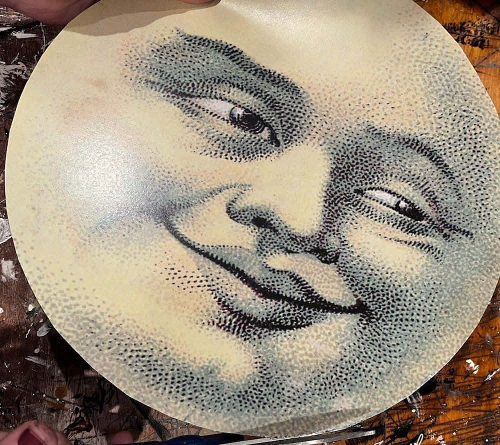 Moon Face 桌布 壁紙 — 一個圓形點陣肖像，描繪一個微笑、閉著眼睛的臉，位於一個有紋理的表面上，由雙手捧著。