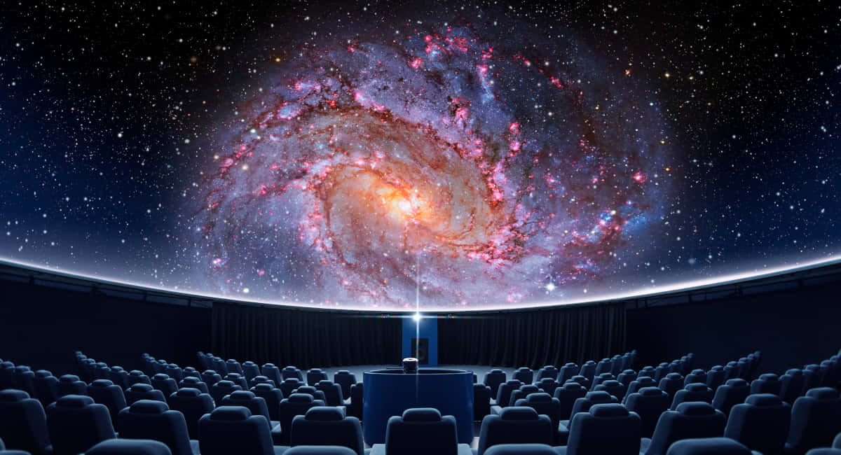 Planetarium 桌布 壁紙 — 一個圓頂劇院展示了一個令人嘆為觀止的螺旋星系，帶有粉紅色和橙色的星雲，背景是繁星點點的夜空。