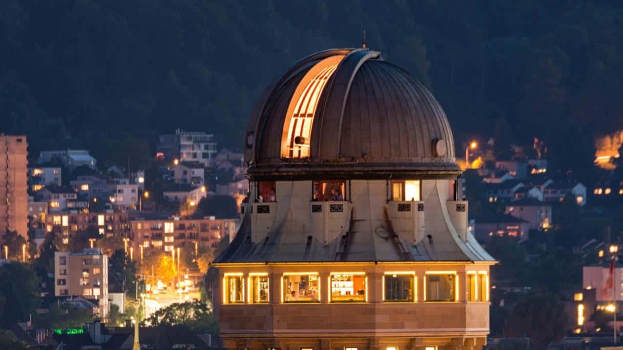 Observatory 桌布 壁紙 — 一個圓頂天文台在暮色中的城市景觀中溫暖地發光，該城市景觀坐落在黑暗的山丘之間。
