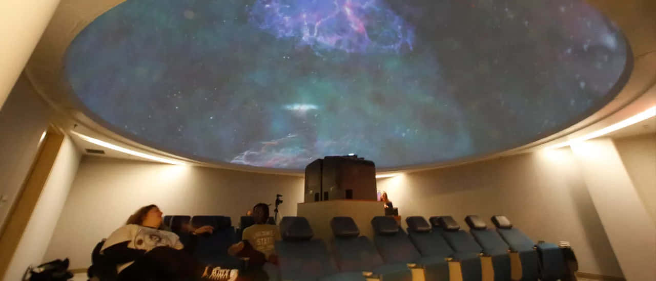 Planetarium 桌布 壁紙 — 一個圓頂天文館，頭頂有星空星雲投影，觀眾坐在藍色椅子的排排座位上。
