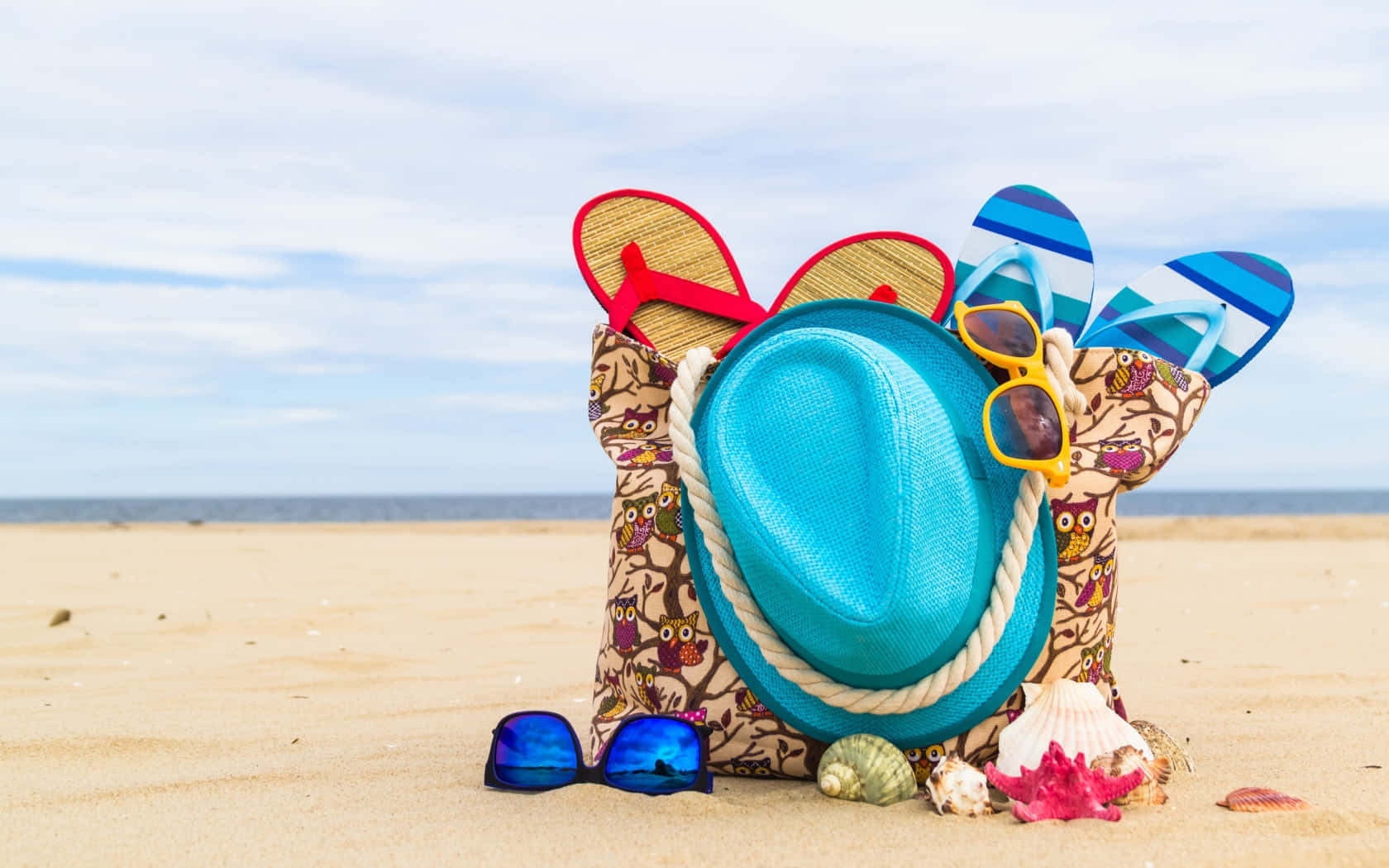 Beach Bag 桌布 壁紙 — 一個圖案沙灘袋裝滿了人字拖、太陽眼鏡和一頂藍色帽子，放在金色沙灘上，旁邊有貝殼，天空多雲。