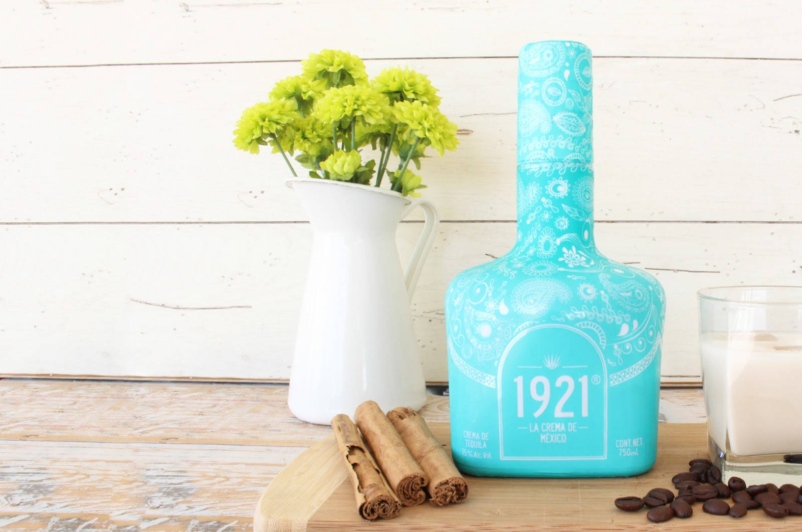 drinks 桌布 壁紙 — 一個土耳其藍色的1921年龍舌蘭酒瓶（佩斯利花紋）放在木板上，旁邊有肉桂棒、可可豆和一瓶綠色花朵。
