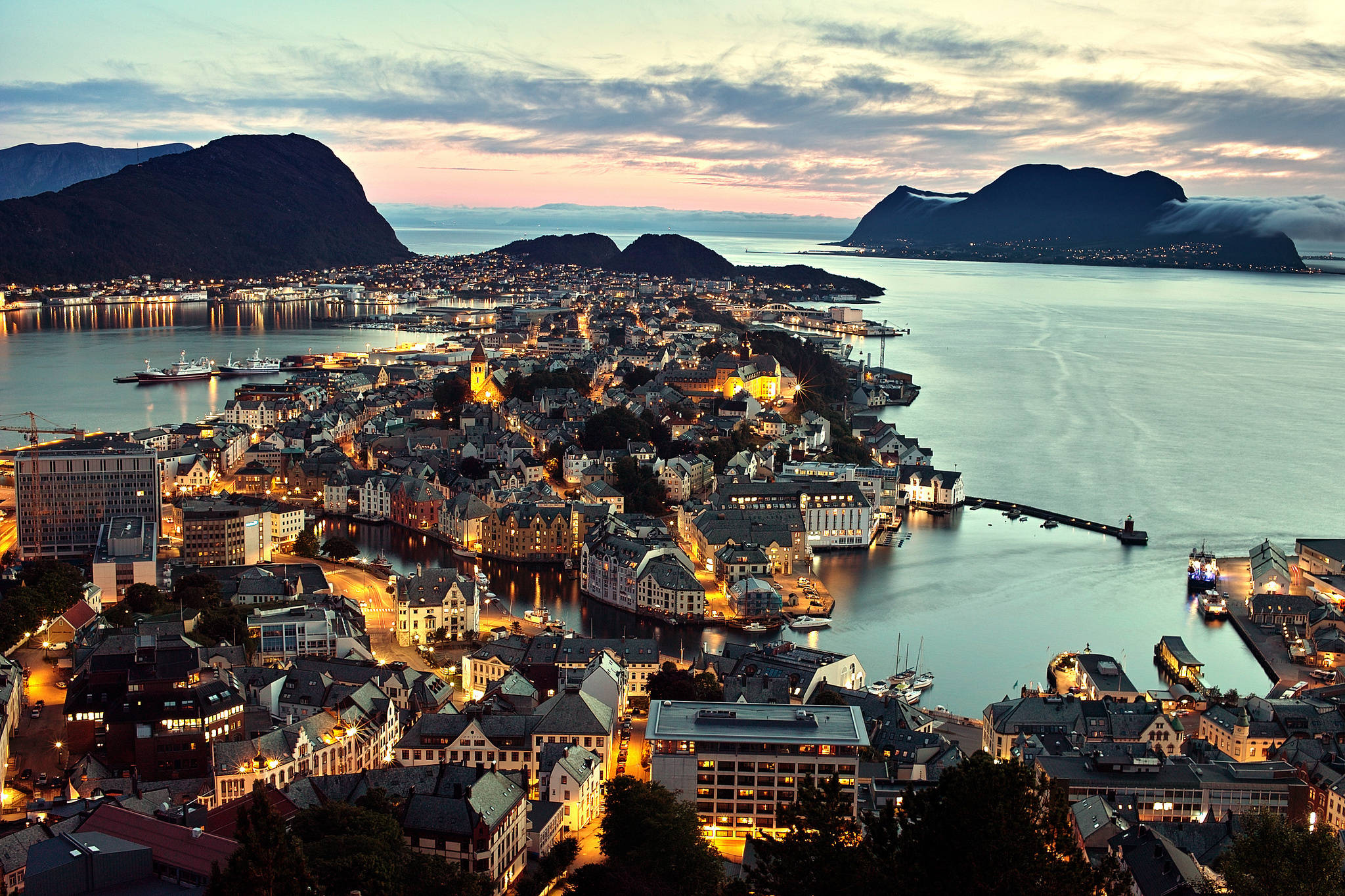 Norway 桌布 壁紙 — 一個坐落在峽灣中的沿海小鎮，在黃昏時分，溫暖的燈光從建築物中閃耀，映襯著柔和的粉紅色和藍色天空。