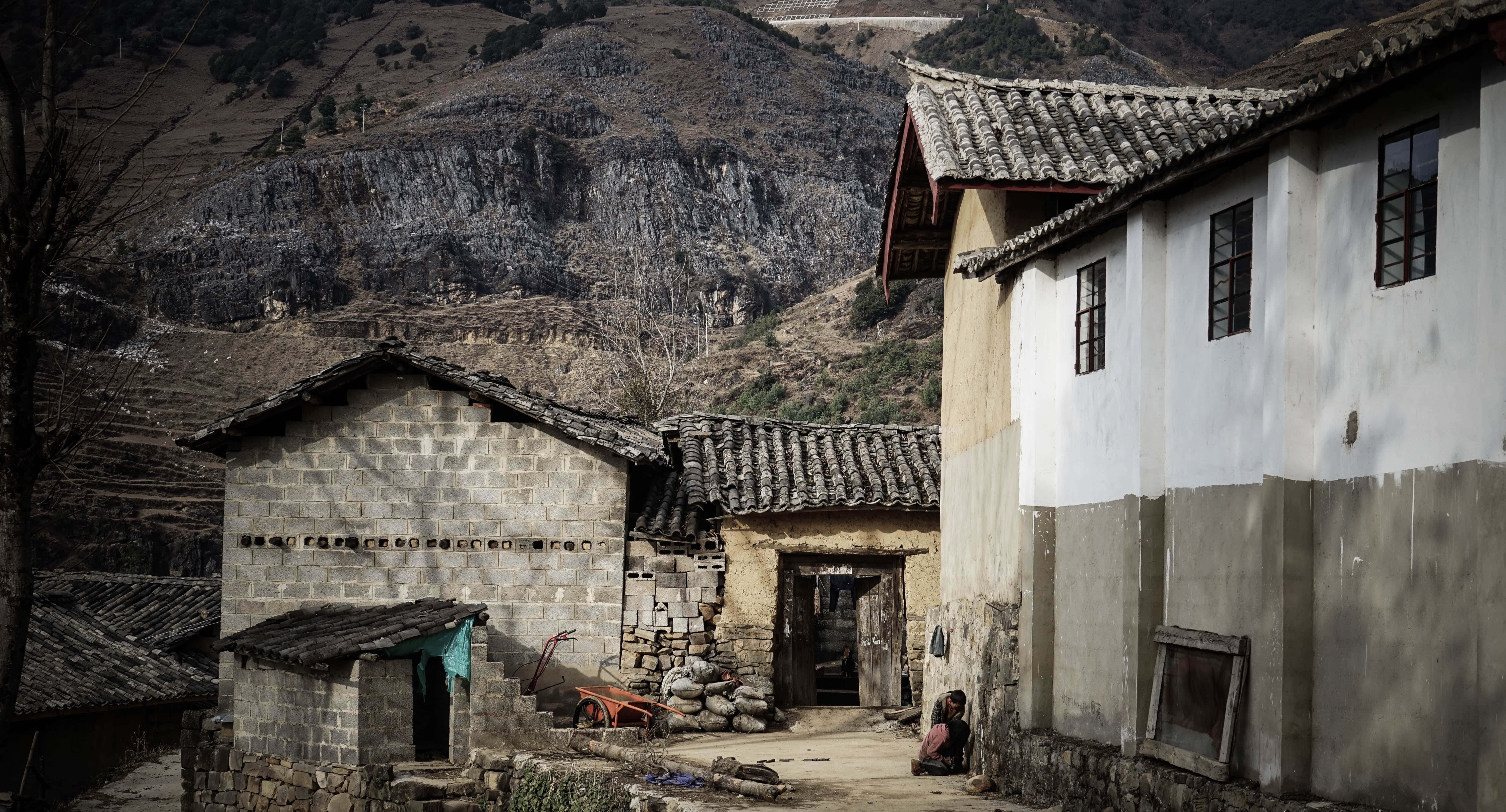 山景 桌布 壁紙 — 一個坐落在崎嶇山脈中的古樸村莊，有風化的石屋和一個人坐在門口附近。
