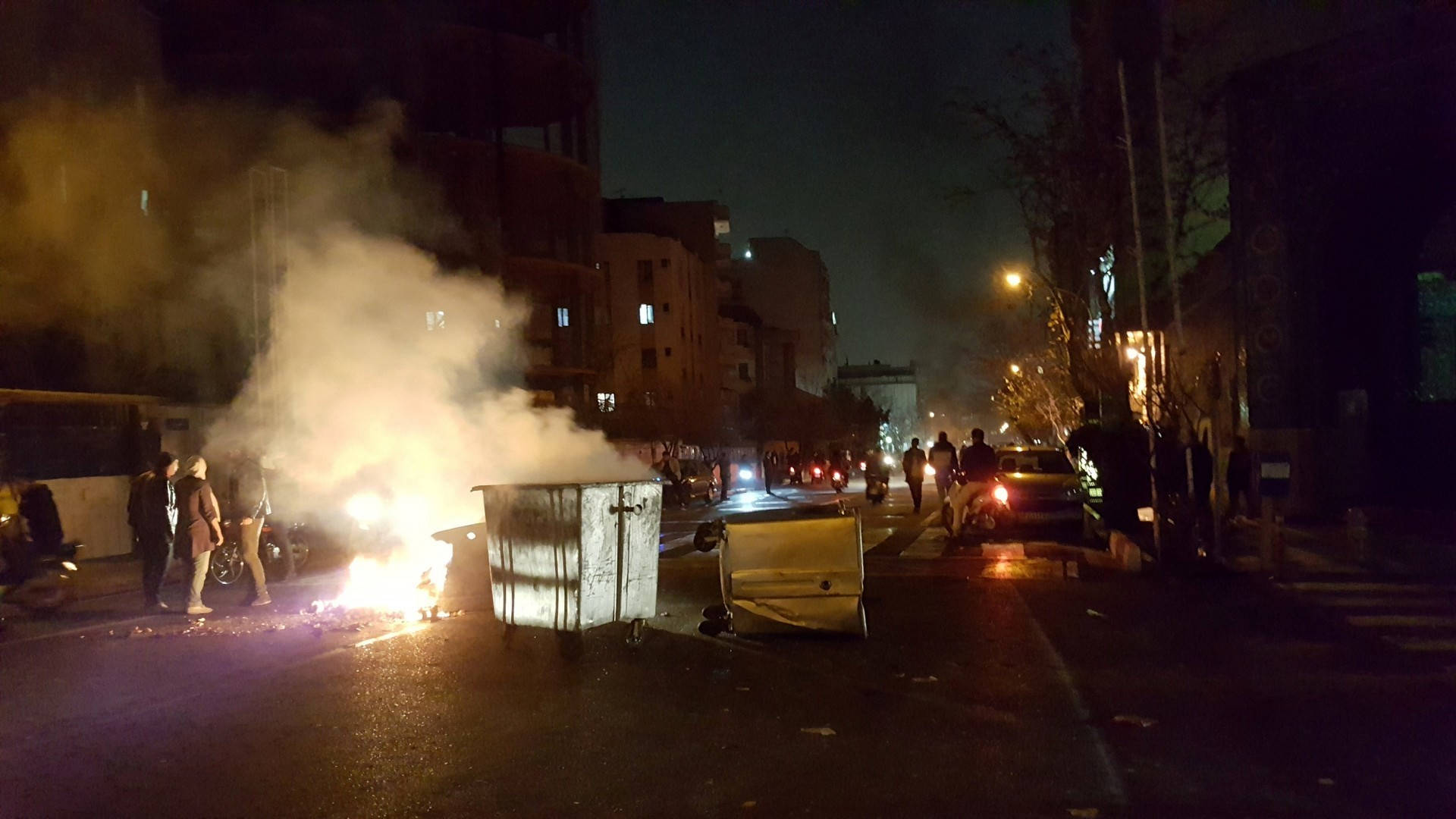德黑蘭 桌布 壁紙 — 一個垃圾箱在夜晚黑暗的城市街道上猛烈燃燒，發出橙色光芒和濃煙，人們聚集在附近。