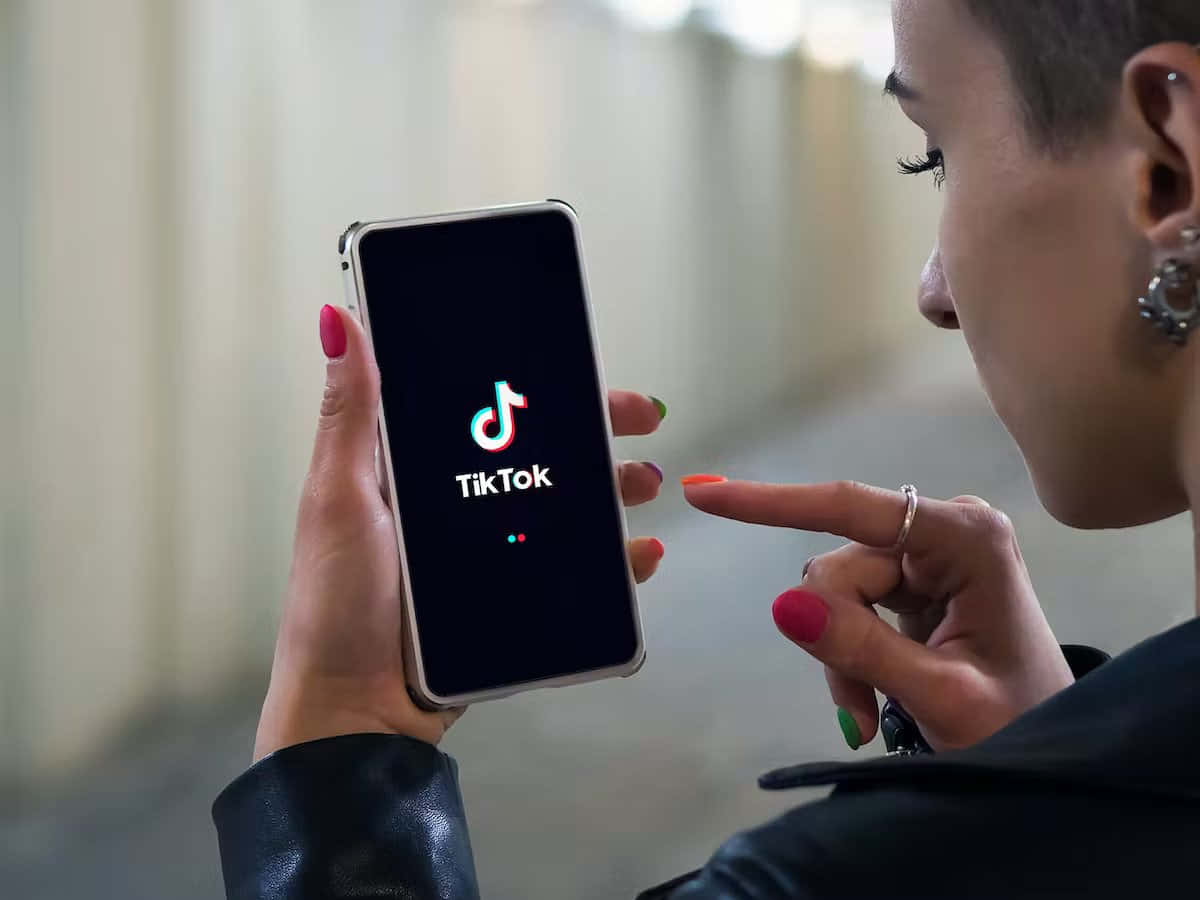 TikTok 動圖 桌布 壁紙 — 一個塗有彩色指甲油的人拿著一部顯示TikTok應用程式標誌的電話，並指向它。