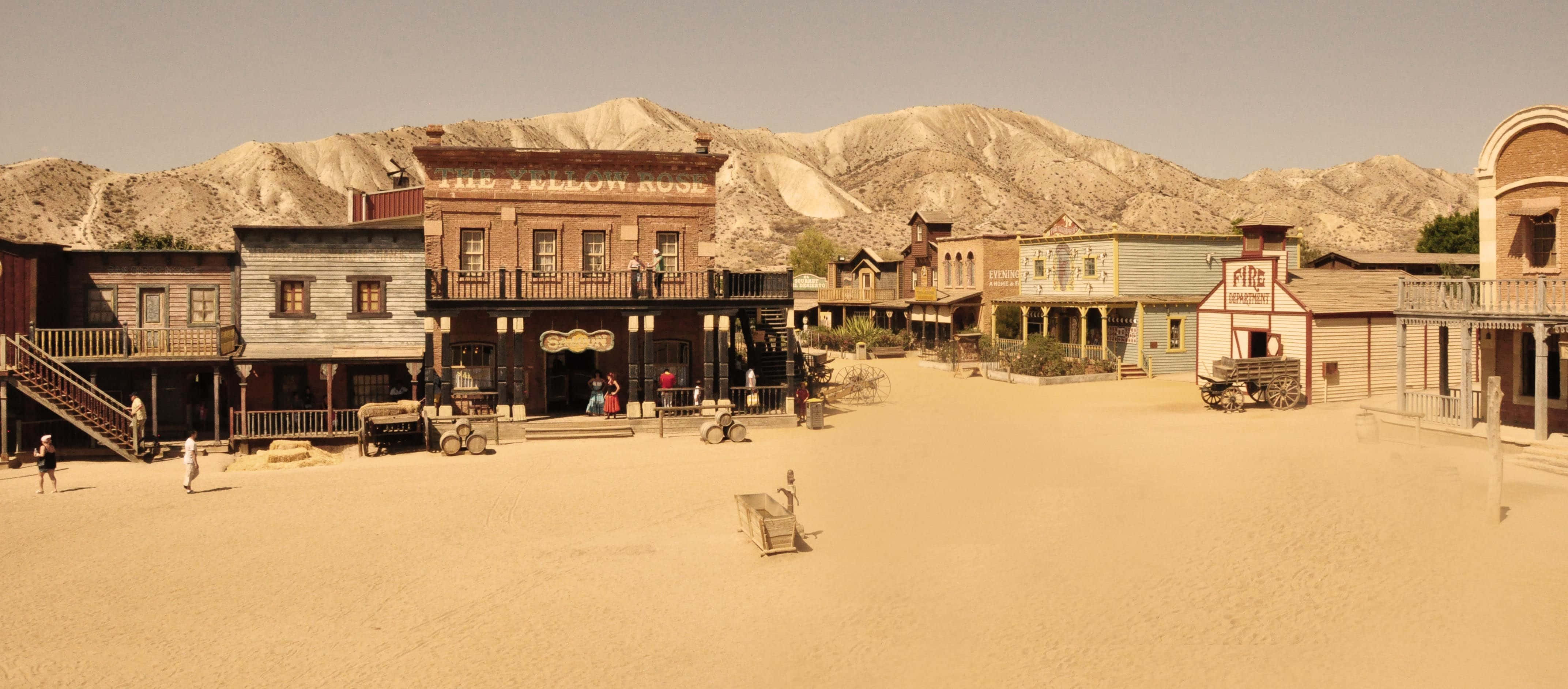 Old Western 桌布 壁紙 — 一個塵土飛揚的西部小鎮廣場，有木製沙龍和背景中的山脈，天空朦朧。