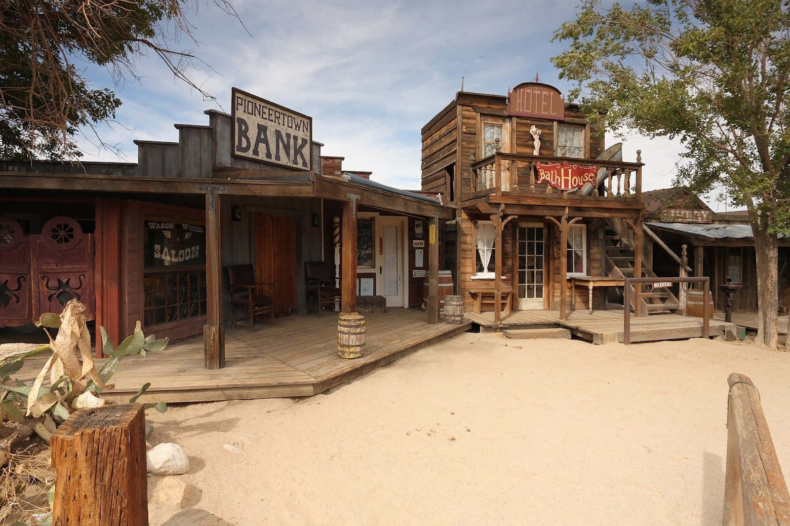 Wild West 桌布 壁紙 — 一個塵土飛揚的西部小鎮廣場，木製建築物上標有銀行和飯店，在明亮的藍天下。