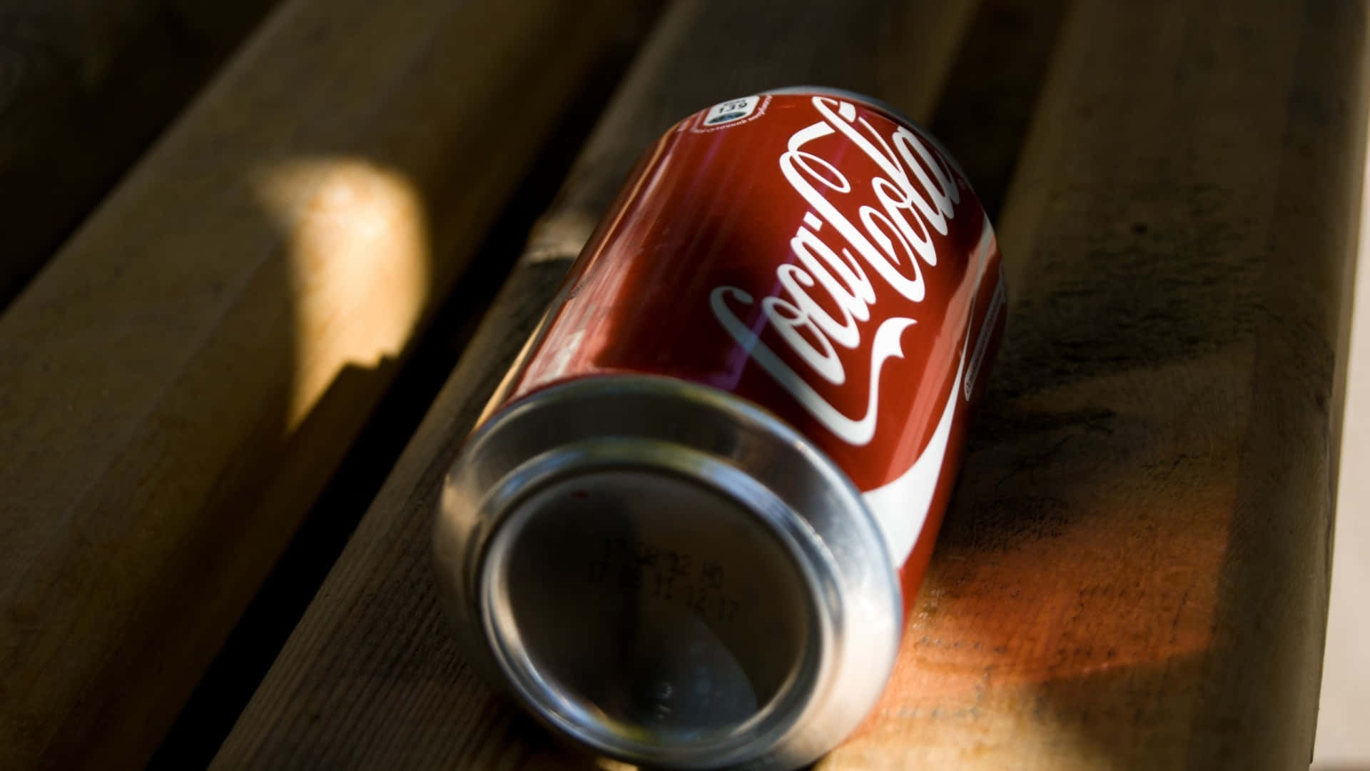 coca cola 桌布 壁紙 — 一個壓扁的可口可樂罐放在飽經風霜的木板上，沐浴在溫暖的陽光中，投下柔和的陰影。
