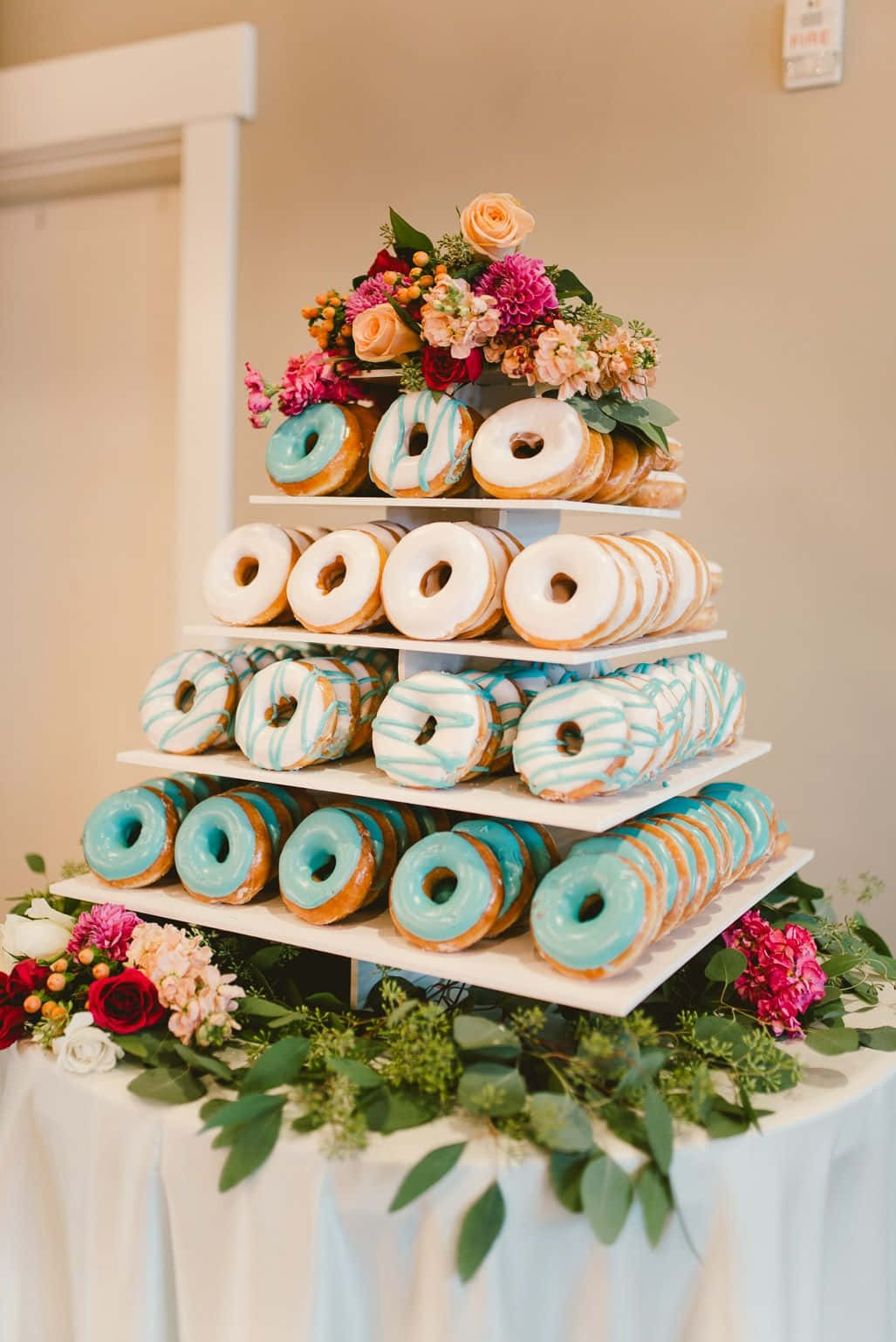 doughnut 桌布 壁紙 — 一個多層甜甜圈塔，飾以色彩繽紛的糖霜甜甜圈和鮮豔的花藝佈置，置於白色桌布上。