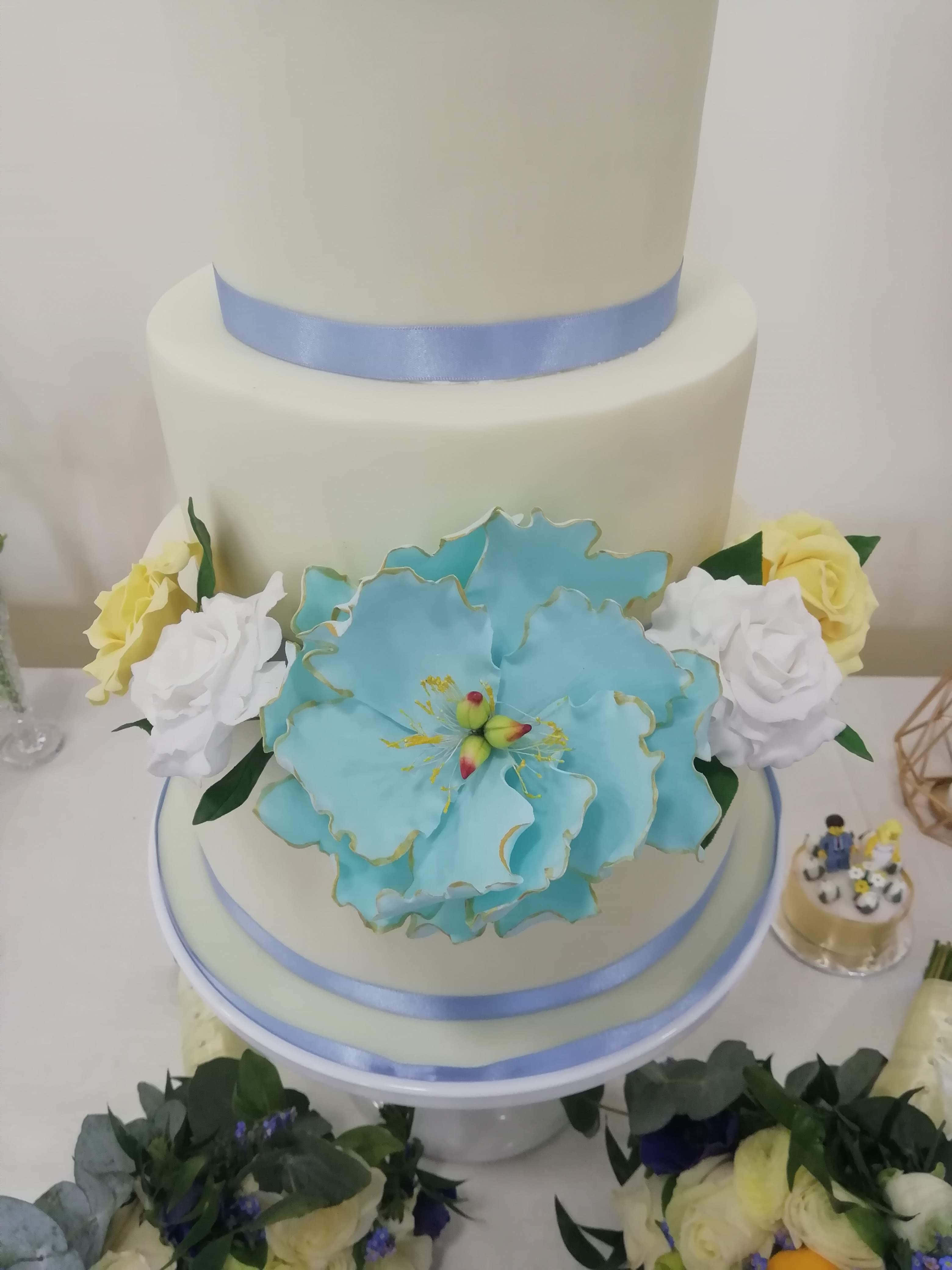 Cake Fondant 桌布 壁紙 — 一個多層白色結婚蛋糕，上面裝飾著一朵大的綠松石糖花和黃白玫瑰，放在白色桌子上。