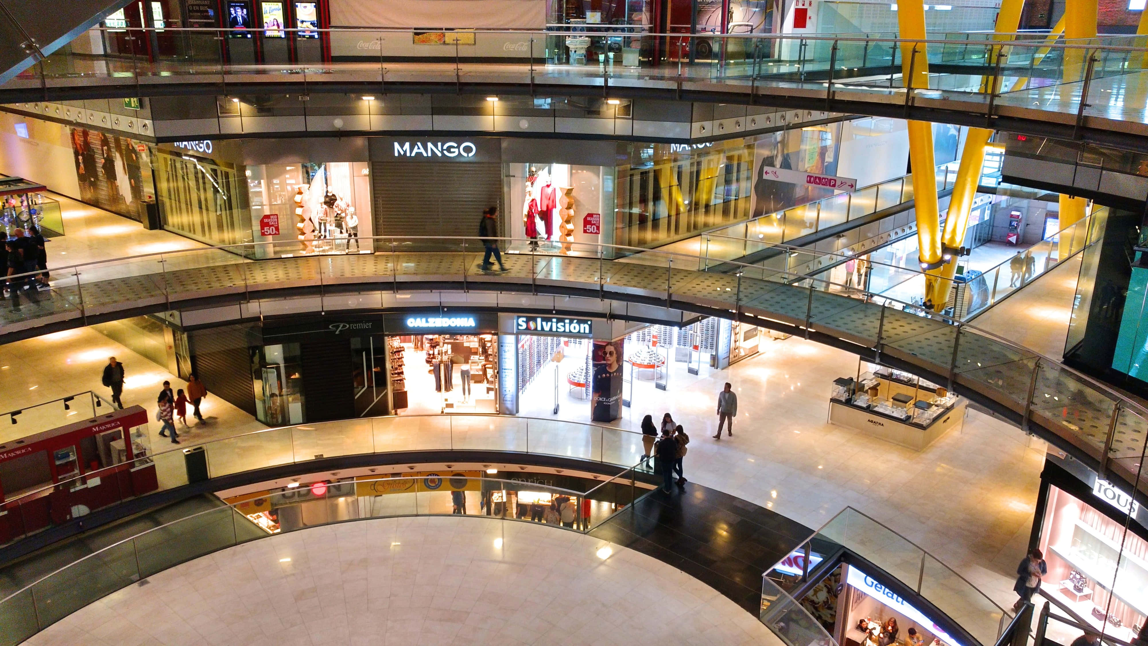 Shopping Mall 桌布 壁紙 — 一個多層購物中心的高角度視圖，設有玻璃欄杆、購物者以及可見的商店標誌，如 Mango 和 Salvision。