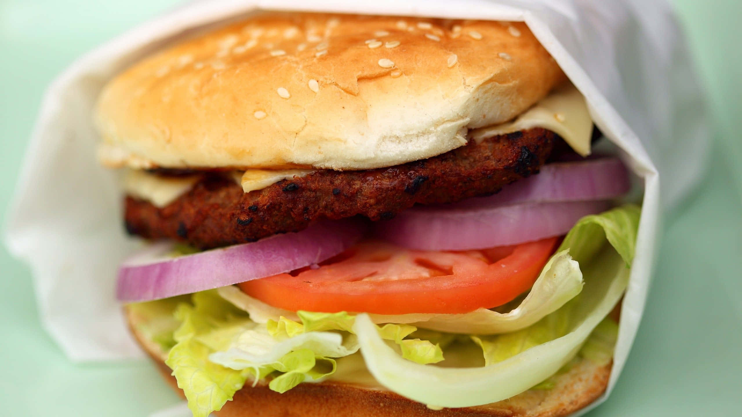 fast food 桌布 壁紙 — 一個多汁漢堡的近距離特寫，配有芝麻麵包、番茄、洋蔥、生菜和融化起司，用紙包裹著。