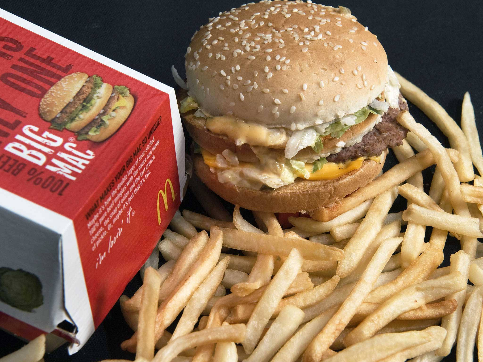 Mcdonalds 桌布 壁紙 — 一個多汁的巨無霸漢堡配芝麻麵包，旁邊有一堆金黃色薯條和其標誌性的紅色盒子，放在深色表面上。