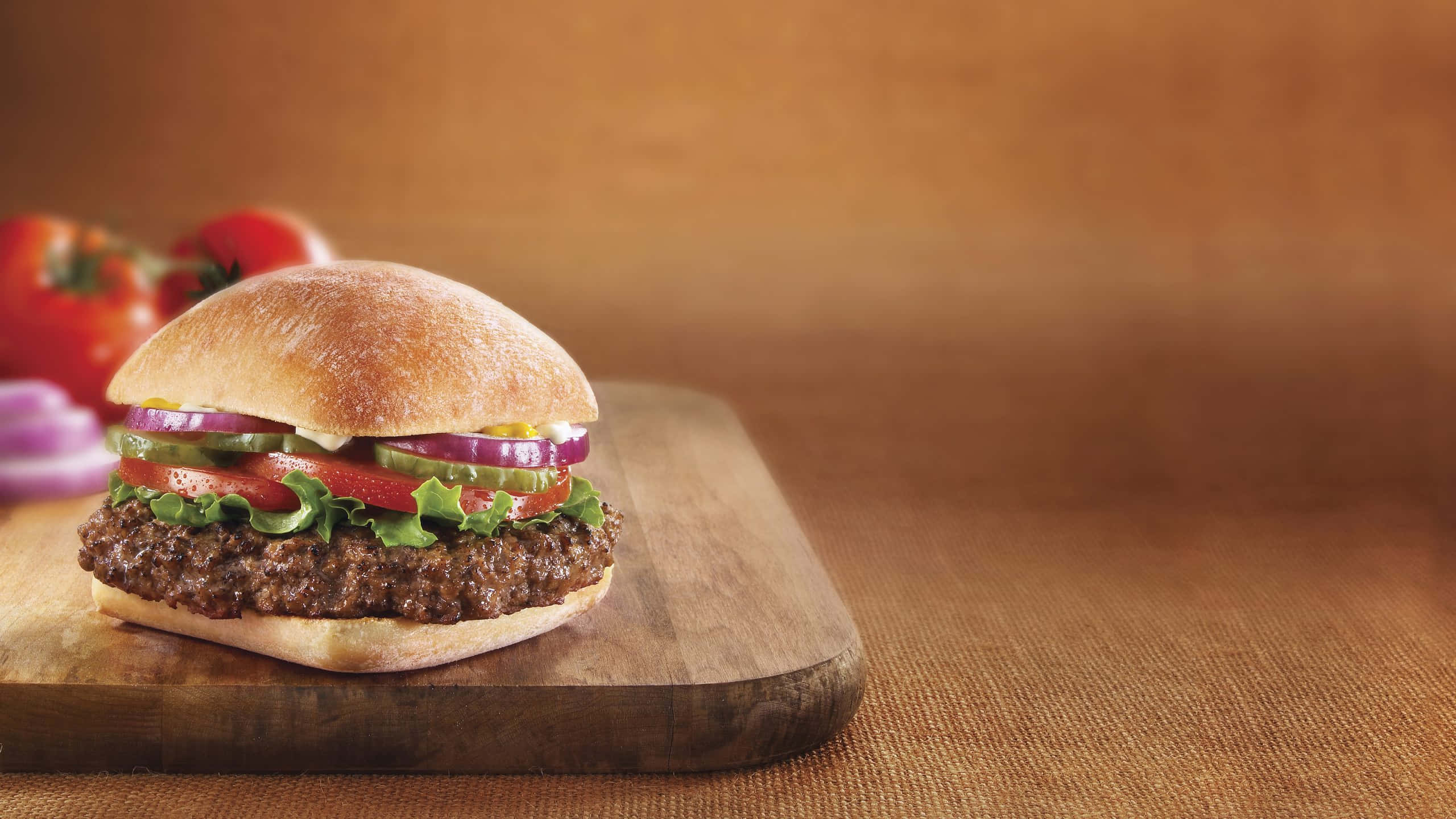 fast food 桌布 壁紙 — 一個多汁的漢堡，配上新鮮生菜、番茄和洋蔥，放在木製砧板上，背景是溫暖的棕色。
