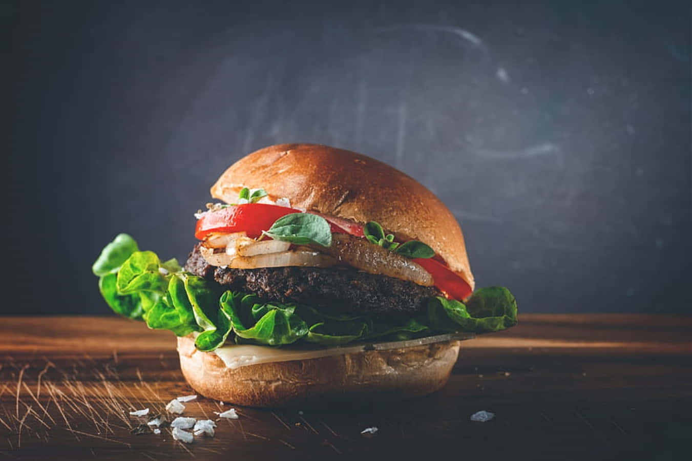 fast food 桌布 壁紙 — 一個多汁的漢堡，配上新鮮的生菜、番茄和洋蔥，放在質樸的木桌上，背景是深色黑板。